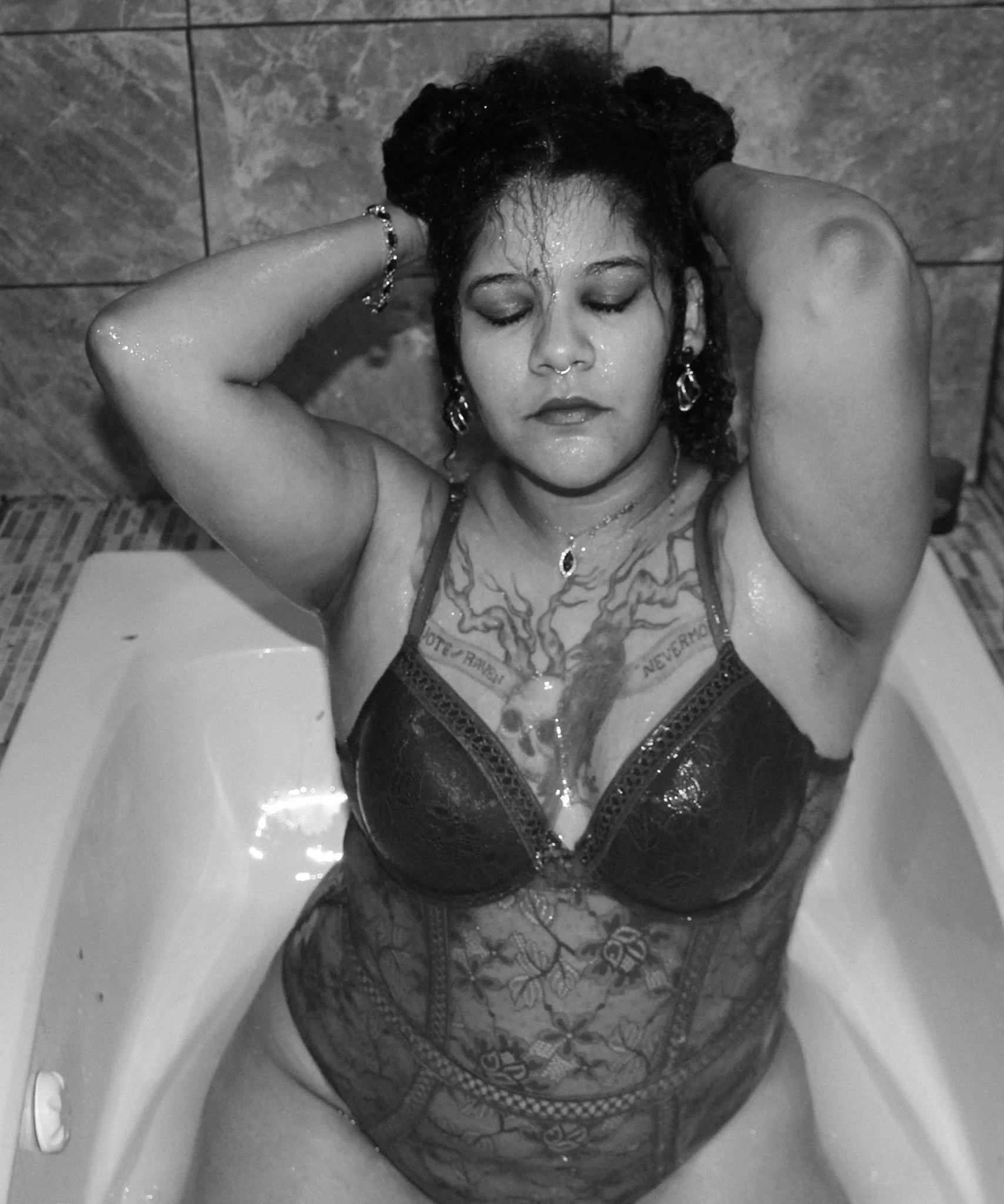 A woman with wet, dark, curly hair reclining with her eyes closed, wearing a lace bodysuit and jewelry, in a bathtub with tiled walls in the background.