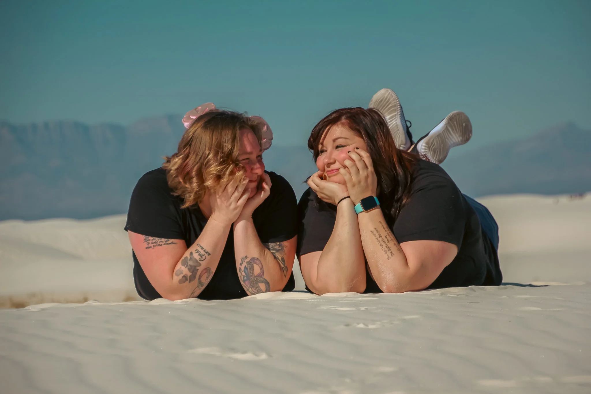 Two women are lying on their stomachs on a white sandy beach, facing each other and smiling. They have tattoos on their arms and are wearing black shirts. The woman on the right has auburn hair and is wearing a smartwatch on her left wrist. They are holding their faces in their hands and appear to be enjoying a joyful moment together.