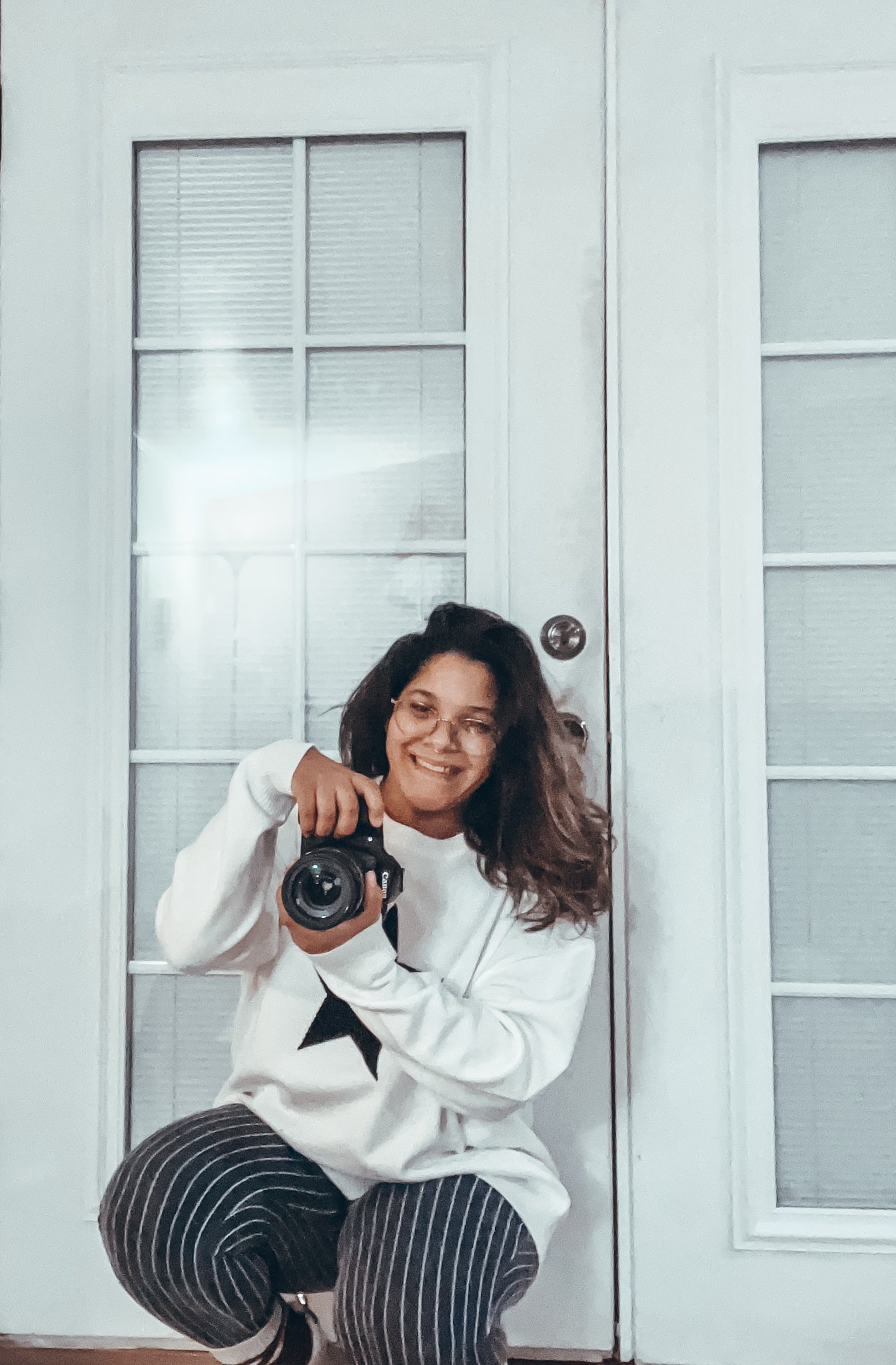 A woman with glasses and long brown hair taking a selfie in a mirror, holding a camera in her right hand, wearing a white jacket, black and white striped pants, and smiling, in front of white door with glass panels.