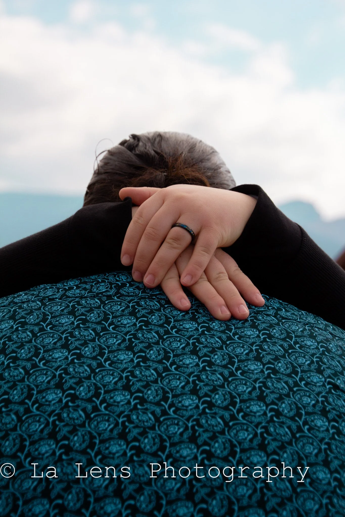 Close-up of two people hugging; one has a black ring on their finger and is wearing a patterned blue and black garment, the other has hair tied back and is wearing a black jacket, outdoors with cloudy sky in background.