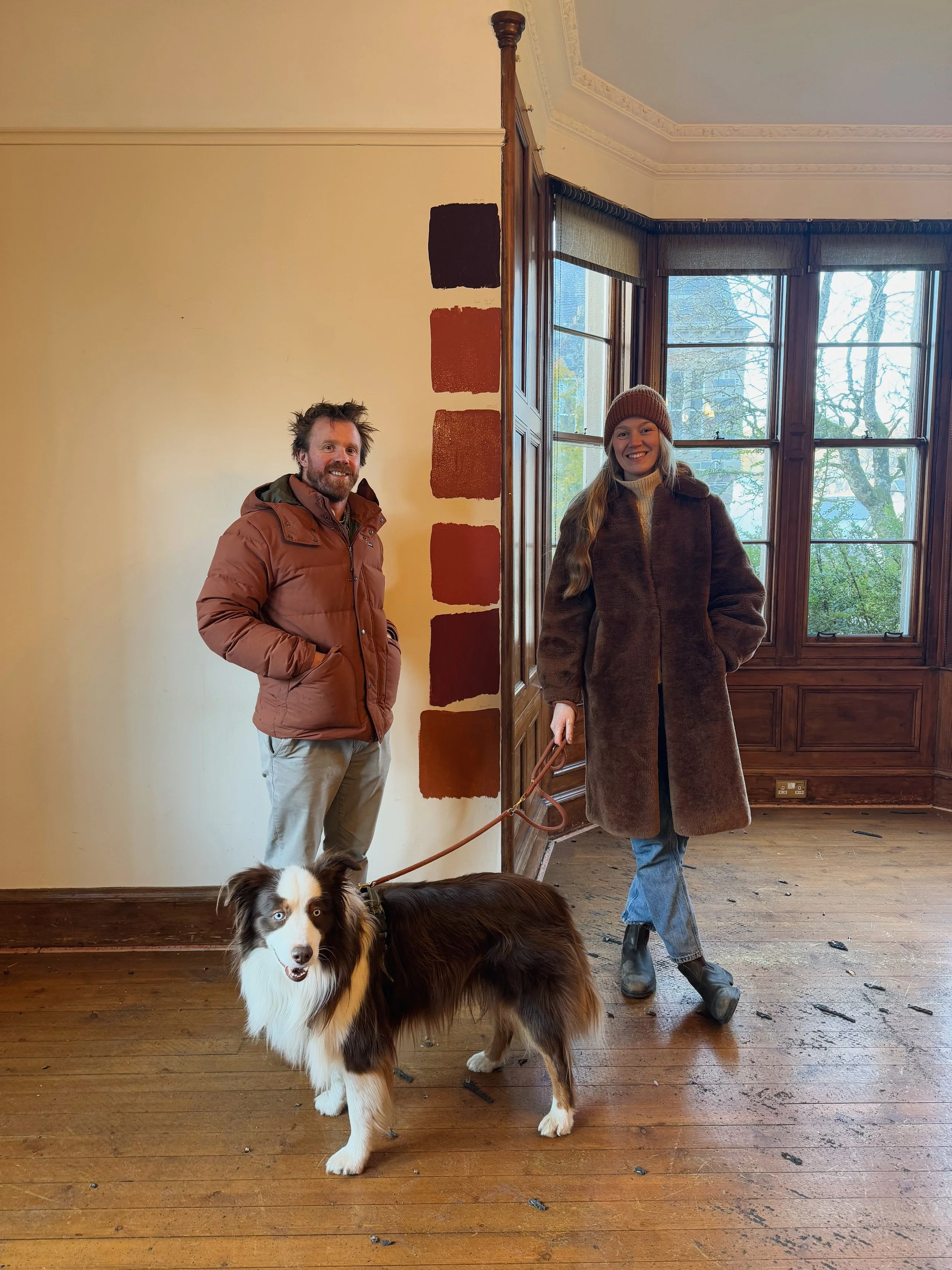 A man and woman standing indoors with a dog. The man is wearing a brown jacket and beige pants, the woman is wearing a dark brown coat and a knit hat, and the dog is an Australian Shepherd with a brown and white coat and blue eyes.