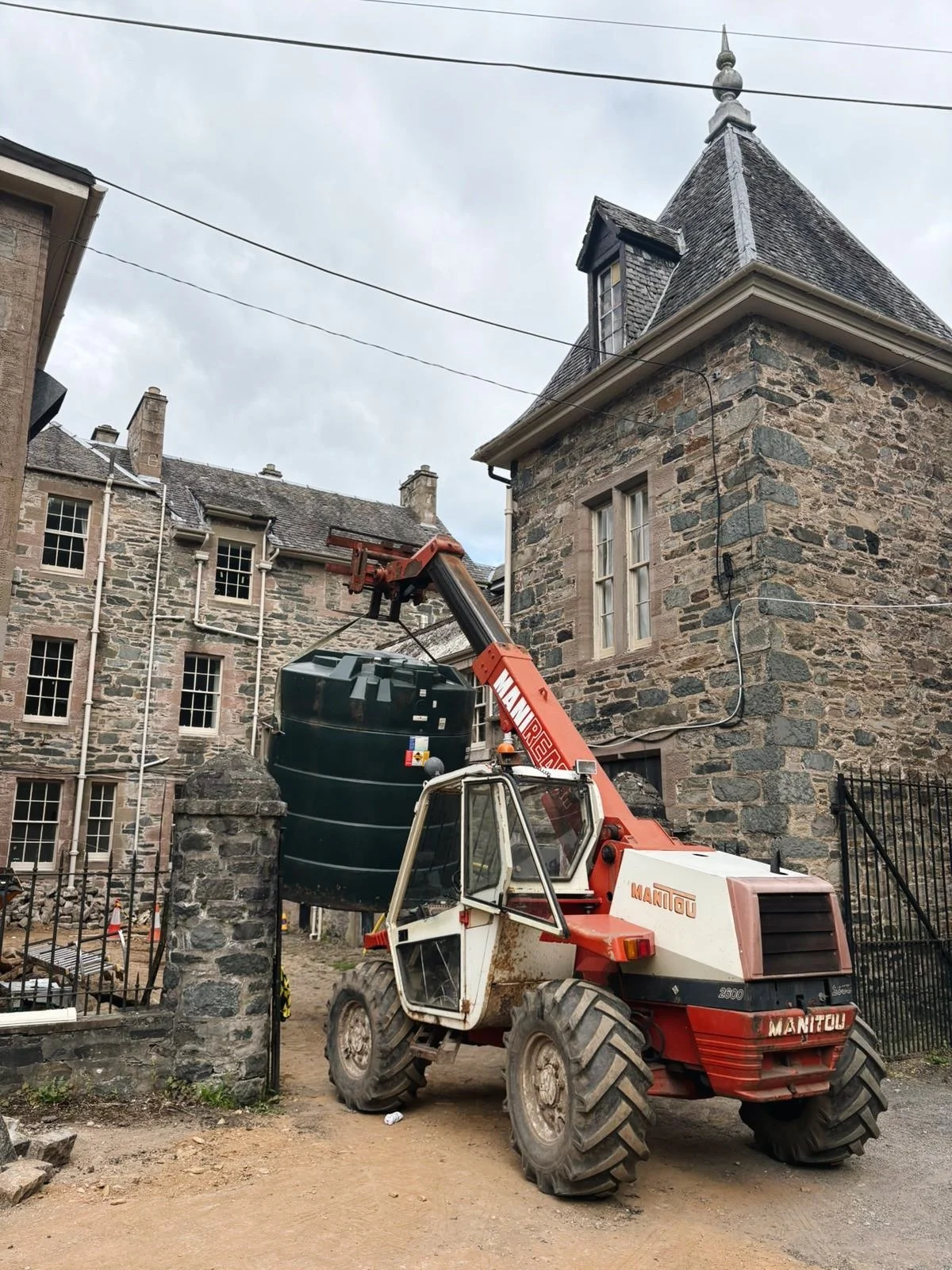 A construction vehicle lifting a large water tank next to a stone building with a turret and multiple windows, on a cloudy day.