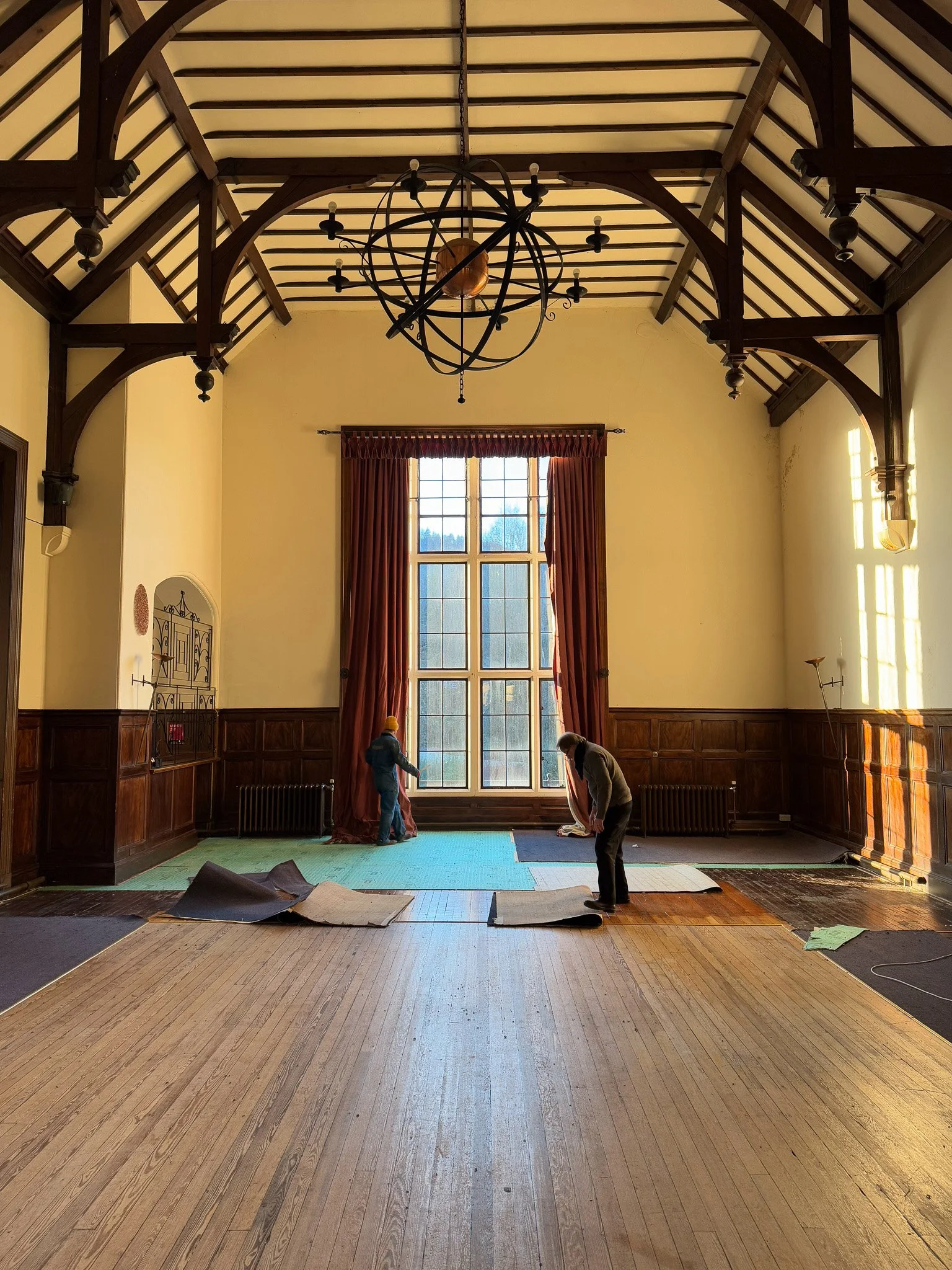  interior  of the birnam hotel baroniall hall room with high vaulted ceiling, large window with curtains, wooden wall paneling, and workers installing or repairing flooring.