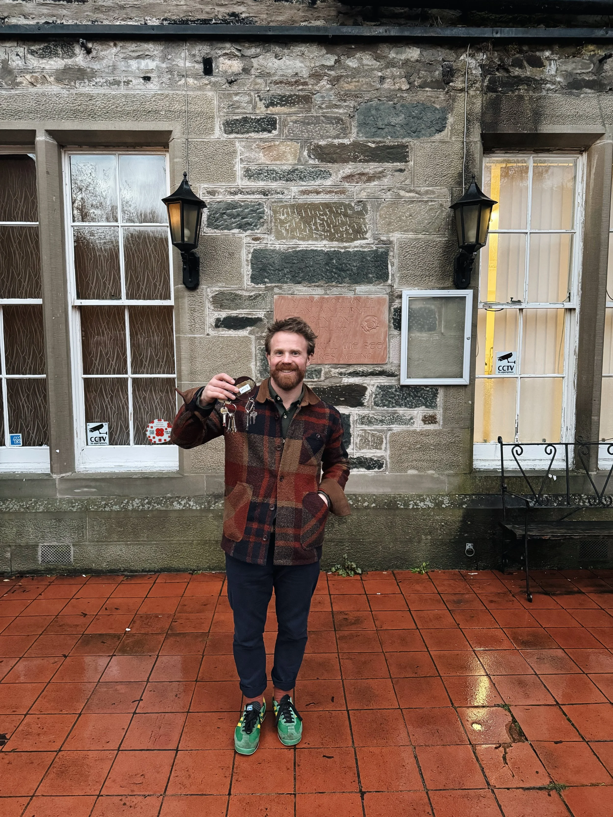 A man with a beard and a smile, wearing a multicolored patchwork jacket, dark pants, and green sneakers, standing outdoors on a wet brick patio holding a set of keys. Behind him is an old stone building with two windows, each with four panes, and a small metal bench near the right window. The building has two black lantern-style lights mounted on the wall and a commemorative plaque partially visible above the man's head.