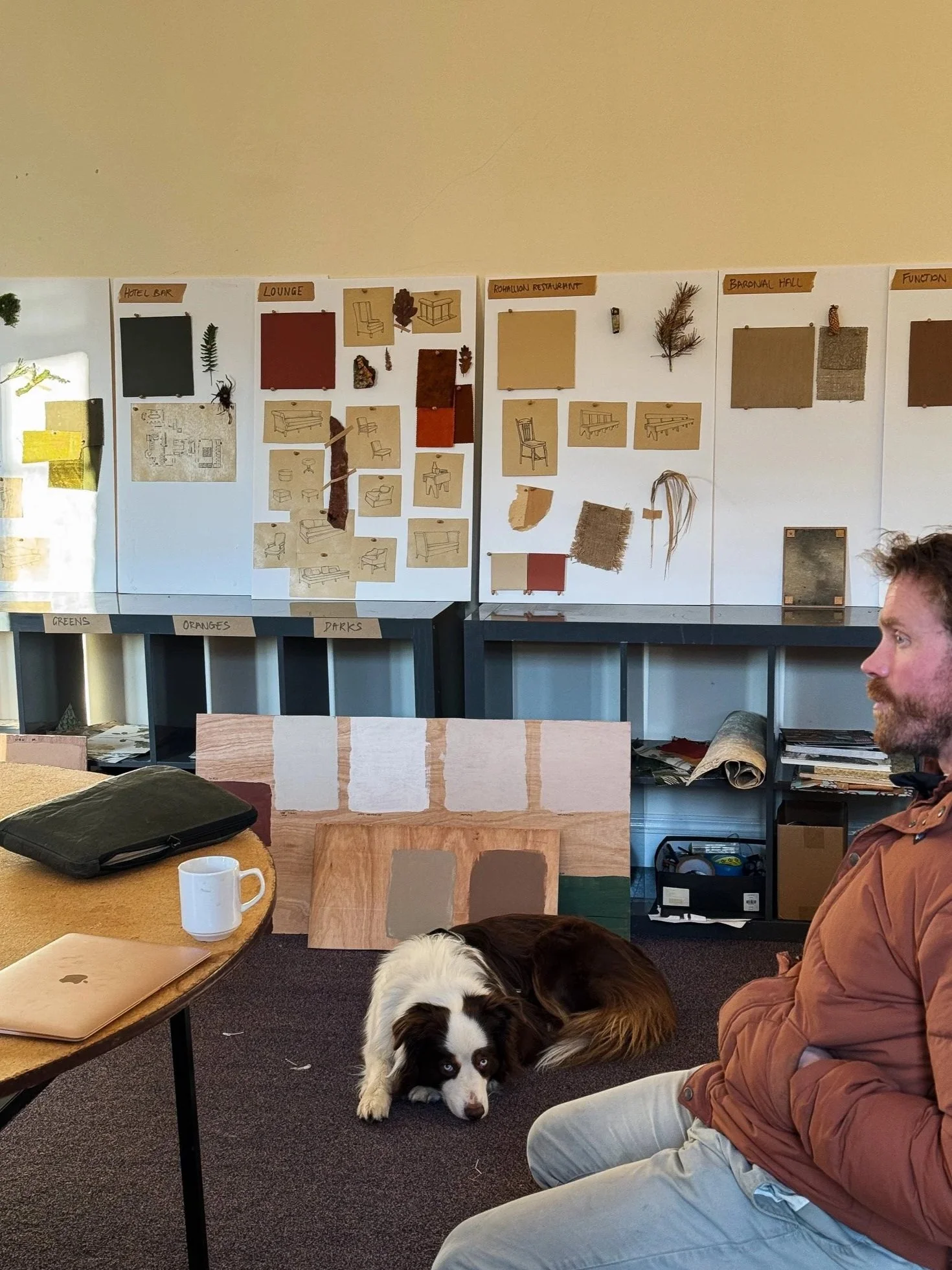 A man sitting indoors next to a Border Collie dog lying on the carpet; behind them, a wall with design boards displaying furniture and room concepts, and a table with a laptop, mug, and backpack.
