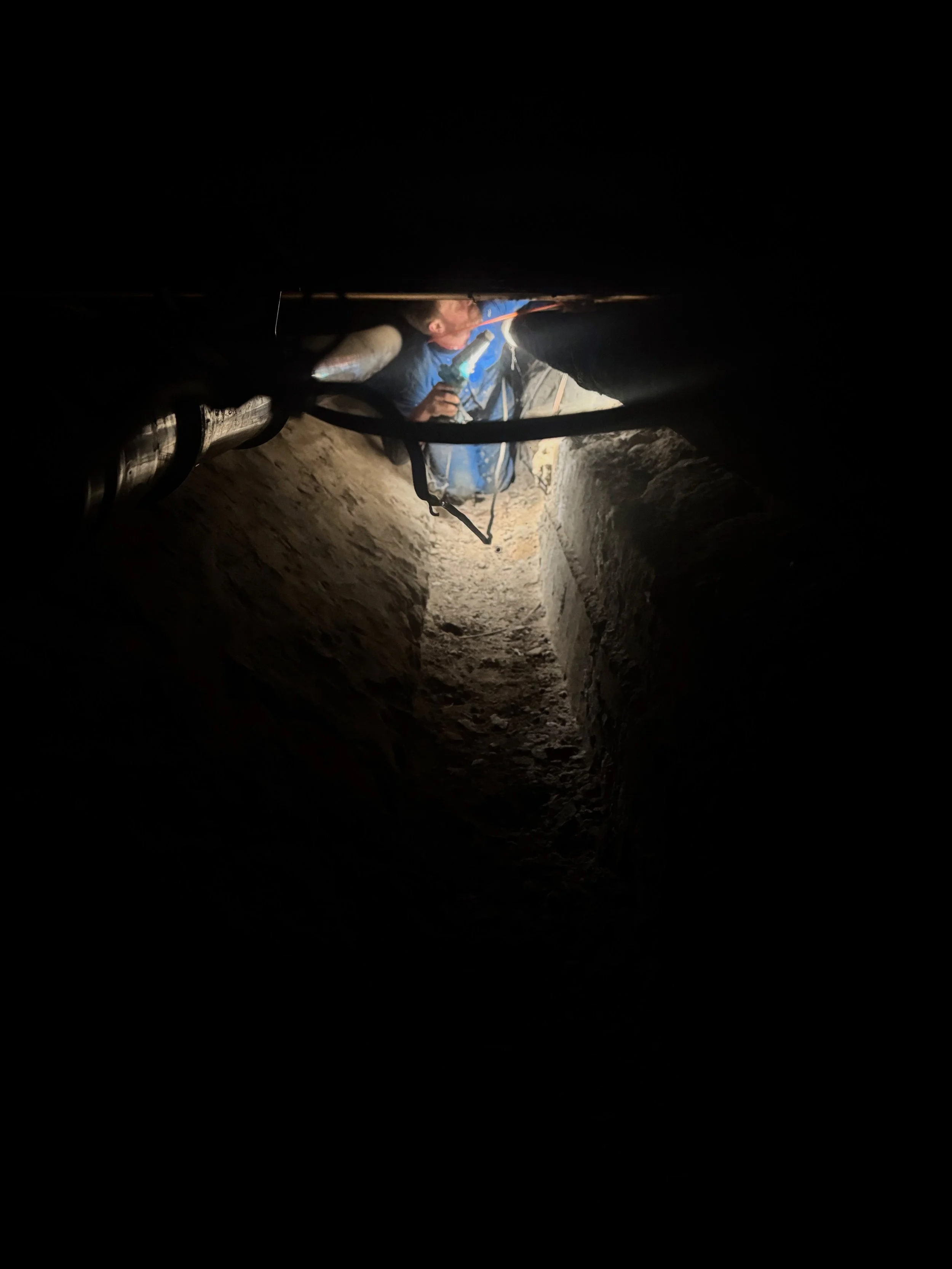 A person with a blue backpack and a water bottle in hand is crawling through a narrow, dark cave tunnel with rough stone walls.