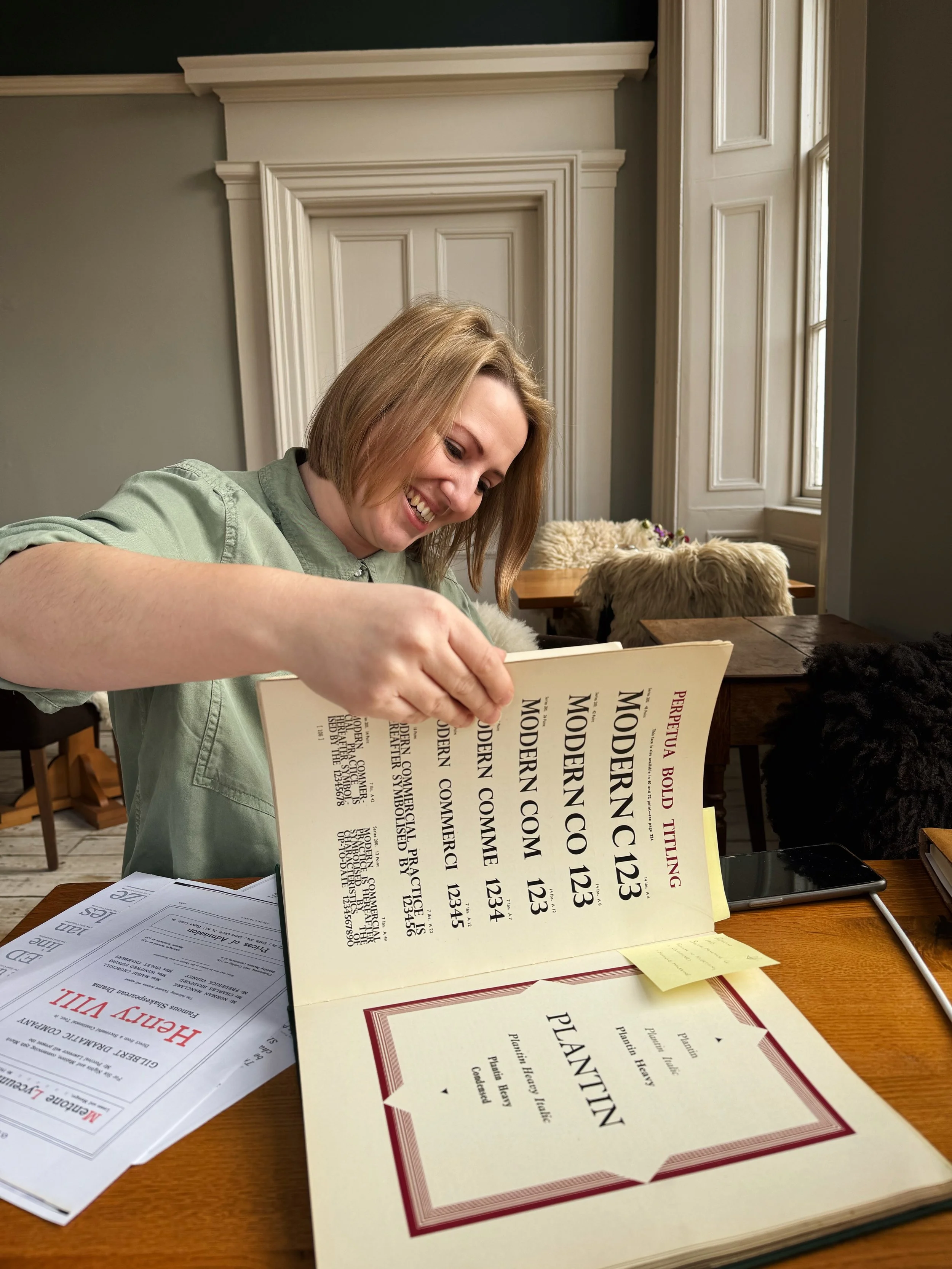 Woman with short blonde hair sitting at a wooden table, flipping through a menu, smiling, with promotional flyers and a tablet on the table, in a room with white walls and a window.