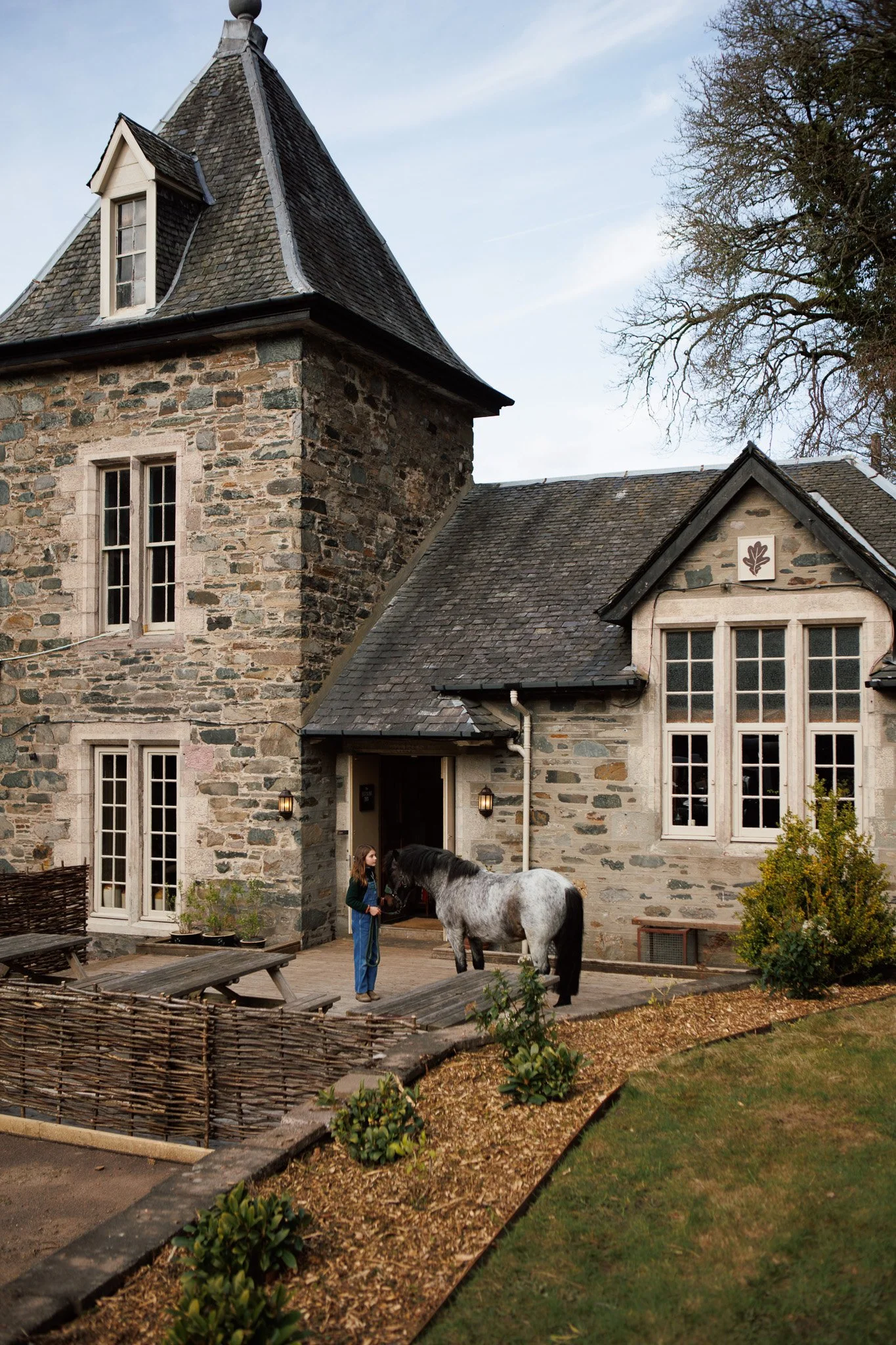 A girl in a blue jacket and jeans standing outside The Birnam Inn pub with a black and white pony.