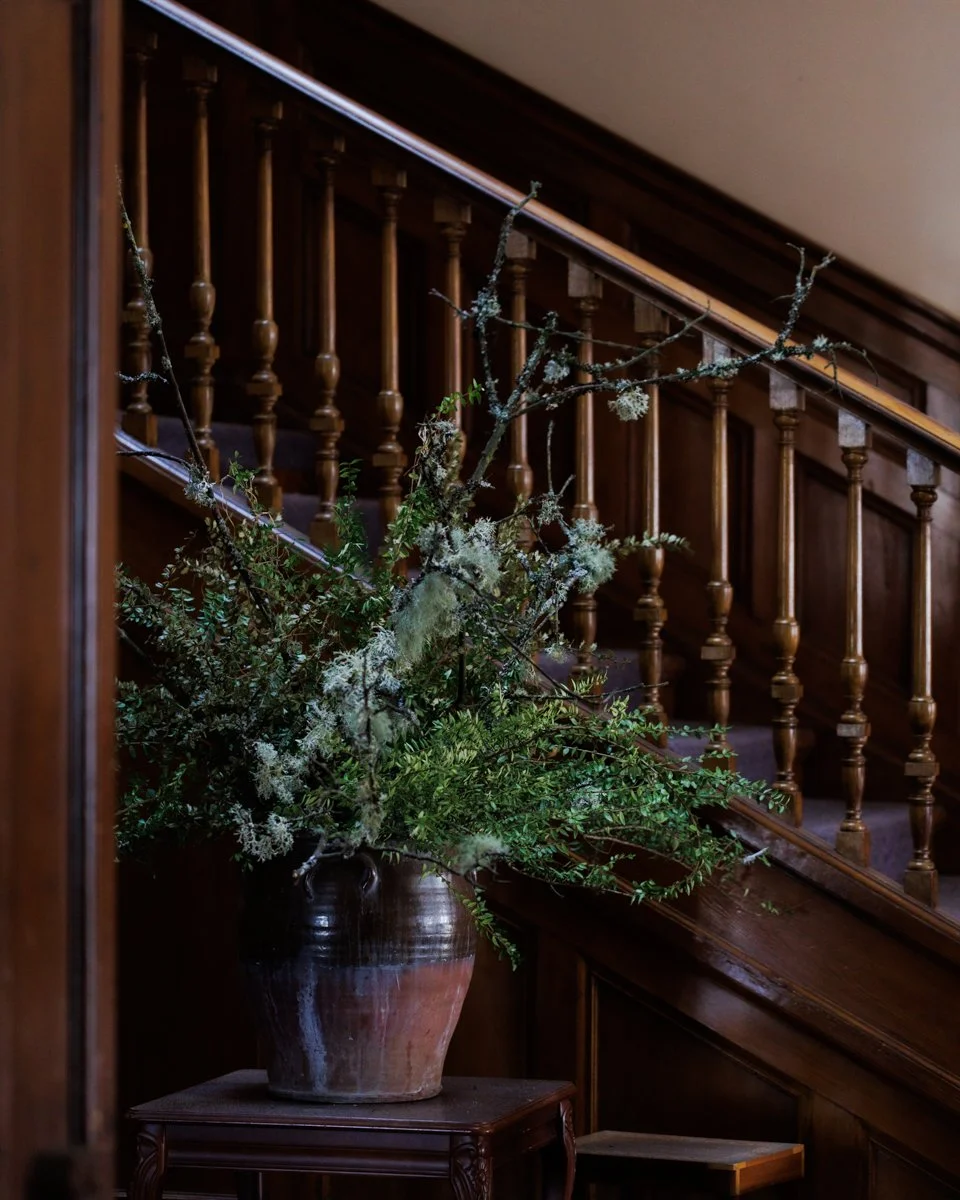A large flower arrangement with branches and greenery in a ceramic vase placed on a small wooden table near a staircase with wooden spindles.