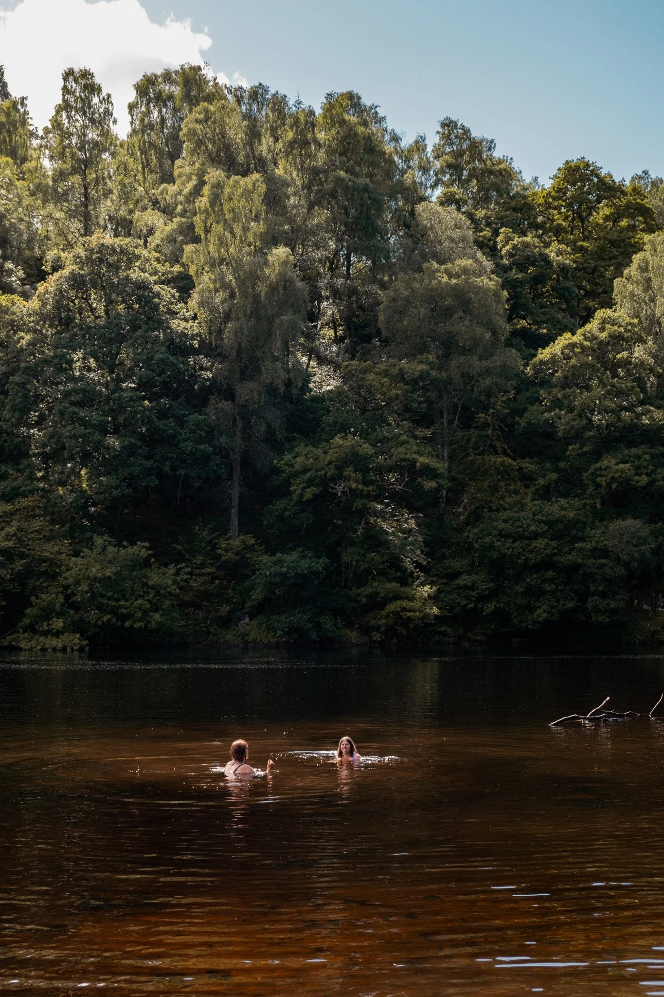 Two people swimming in a dark-colored lake surrounded by lush, green trees under a partly cloudy sky.