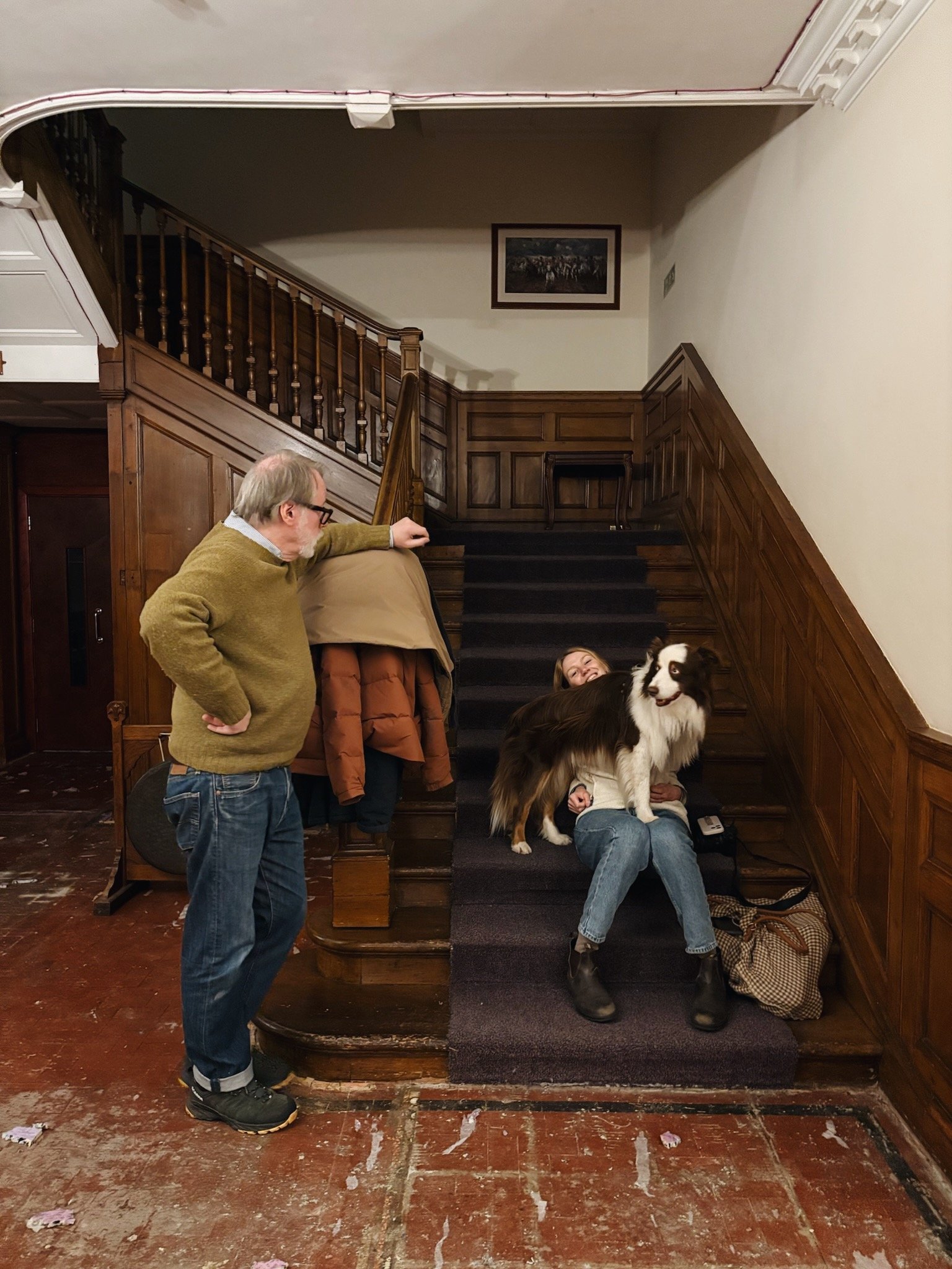 birnam hotel designers kim grant and eric bremner in the lobby with coats hanging on the side of the stairs. The setting appears to be indoors with wooden paneling on the walls and framed artwork above.