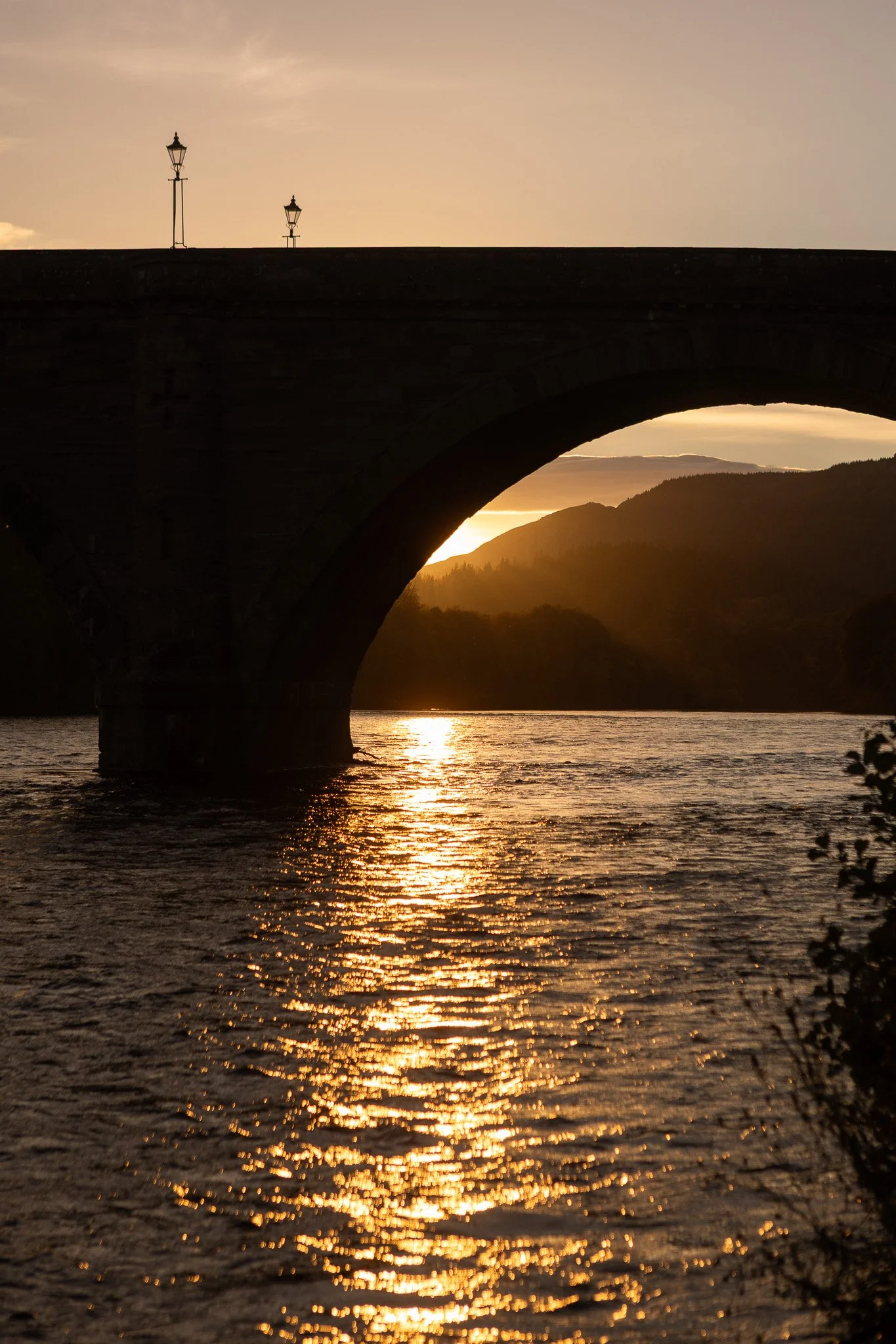 Sunset view of a bridge over a river with mountains in the background.