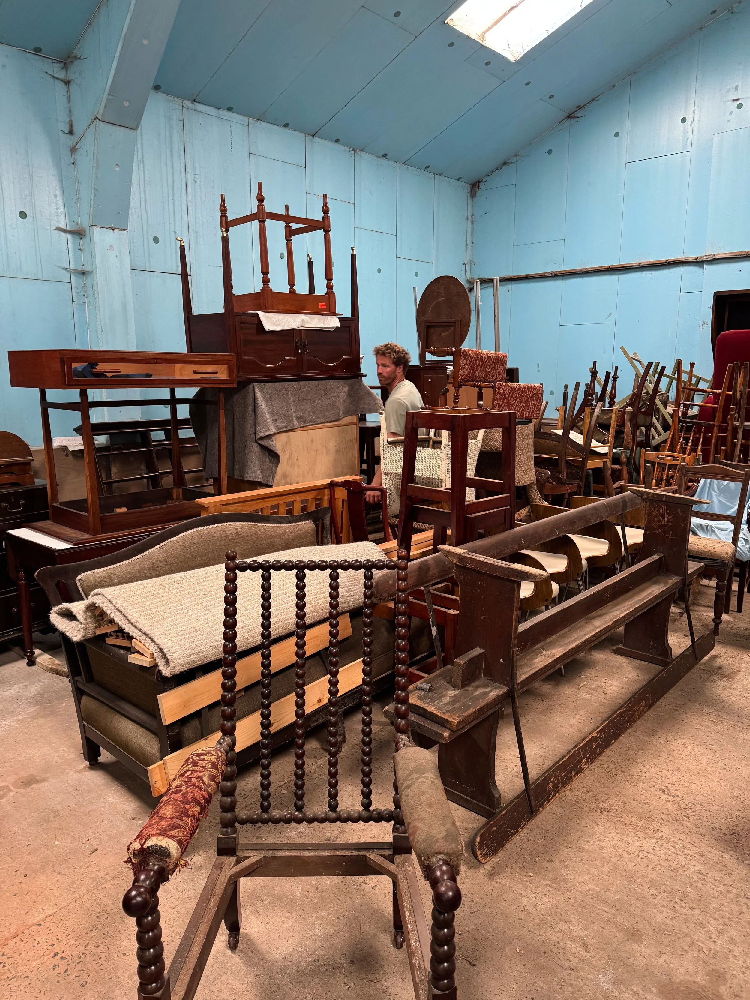 An interior space filled with various antique furniture pieces, including chairs, tables, and benches, in a room with blue walls and a concrete floor.