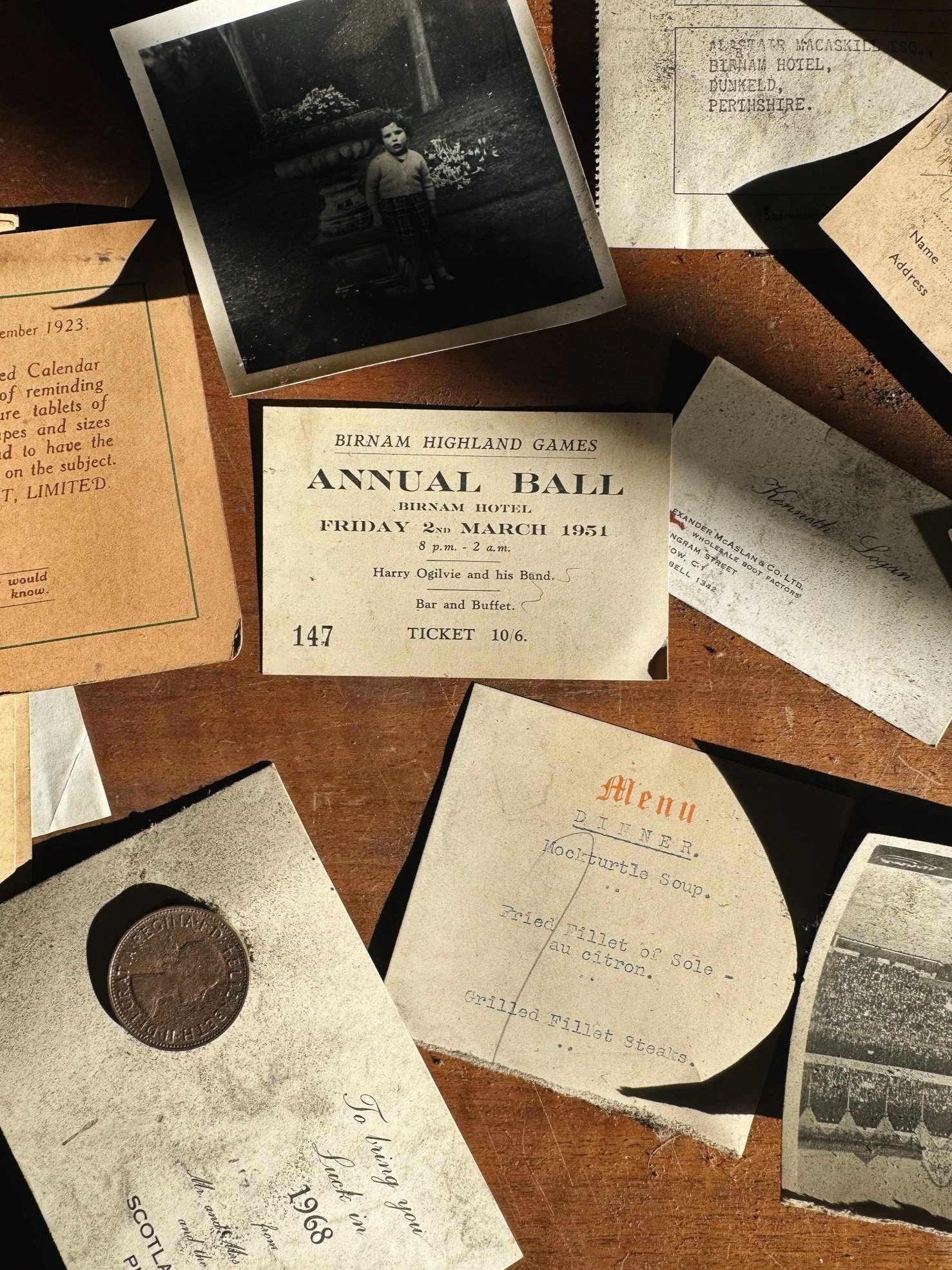 Collection of vintage papers and tickets, including a 1951 Birnam Highland Games annual ball ticket, a menu from a diner, a photograph of a young child in front of a fountain, a coin, and various old handwritten or printed notes scattered on a wooden surface.