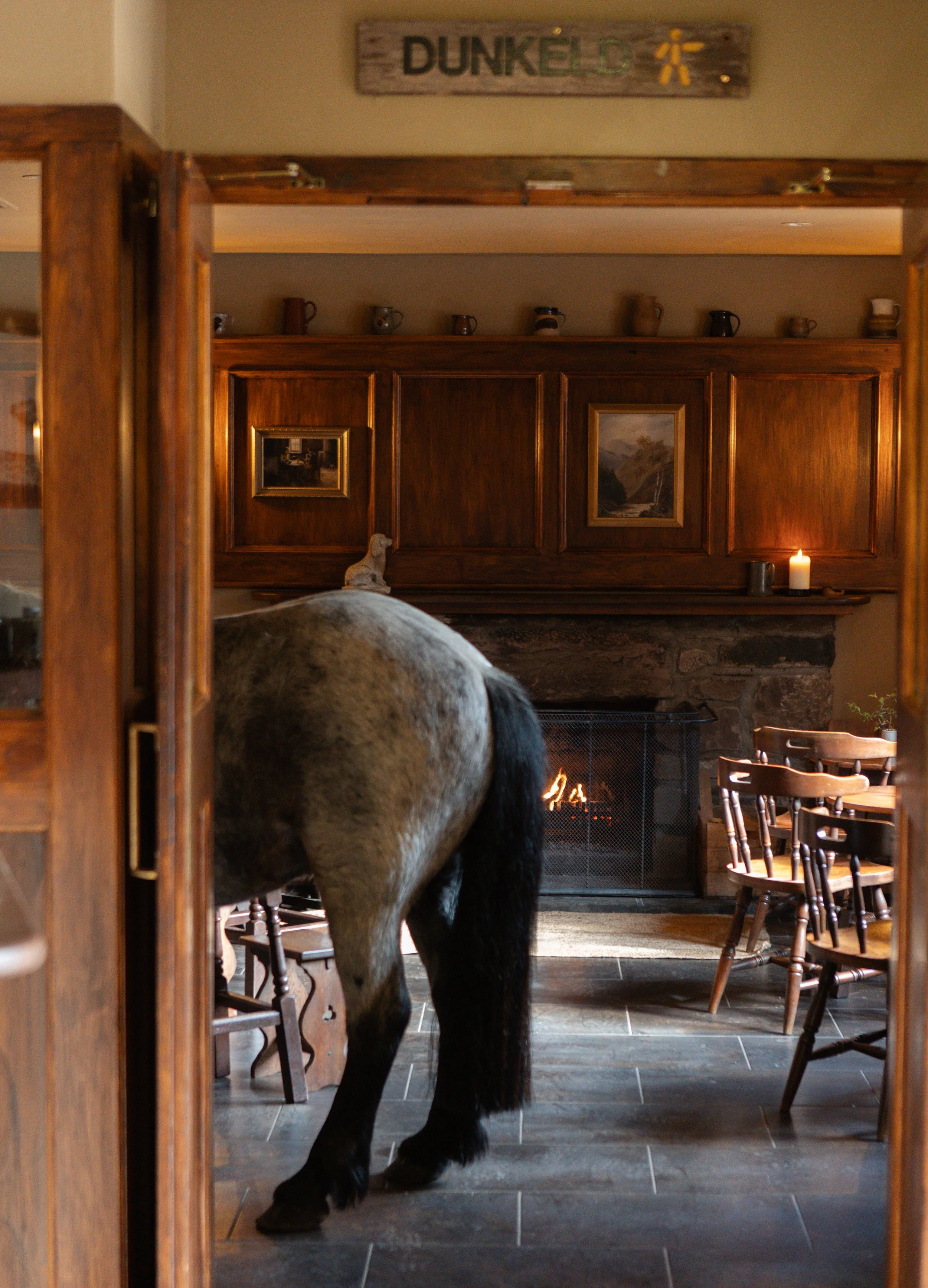 A horse standing in a cosy, rustic pub with wooden chairs, a stone fireplace, and framed landscape paintings, viewed through a wooden doorway with a 'Dunkeld' sign above.