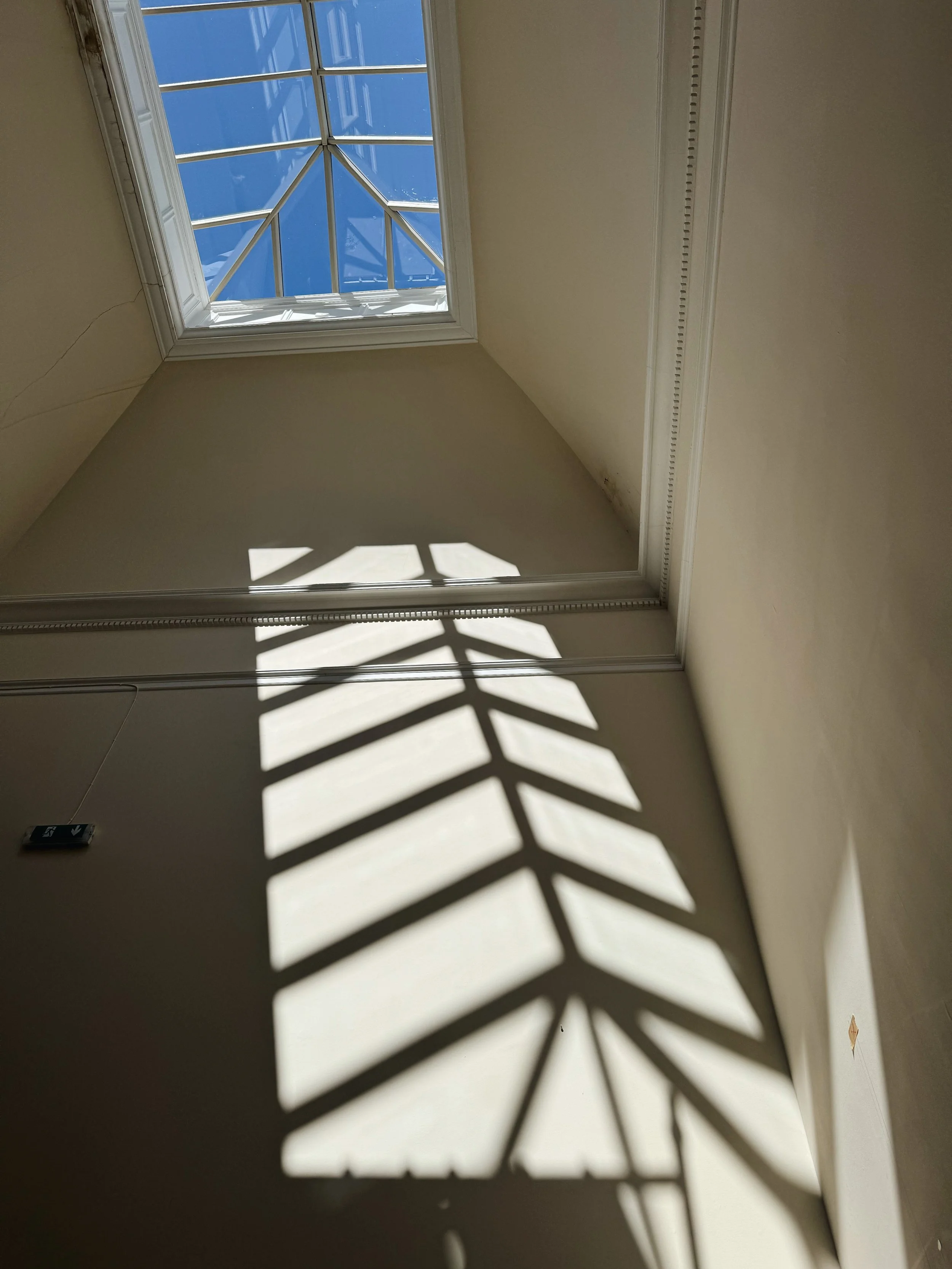 Sunlight casting shadow of a decorative window on a wall inside a room, with a ceiling skylight showing blue sky and a metal framework.