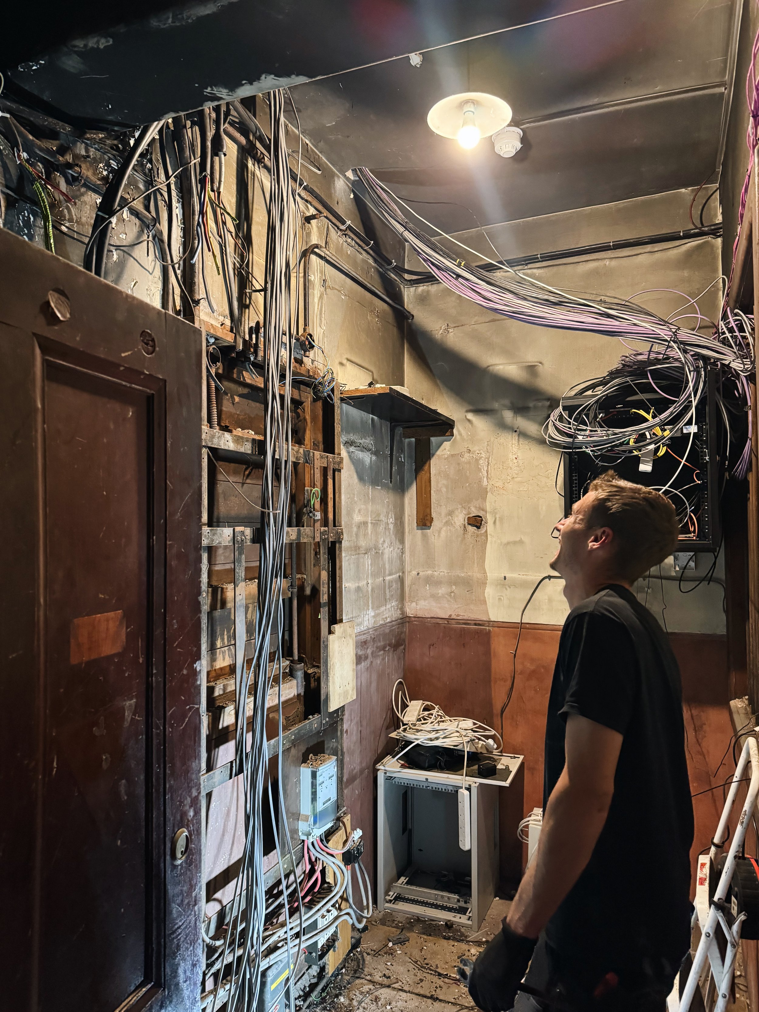 A young man in black shirt and gloves working on a wall with exposed wires and electrical panels in a room under renovation.