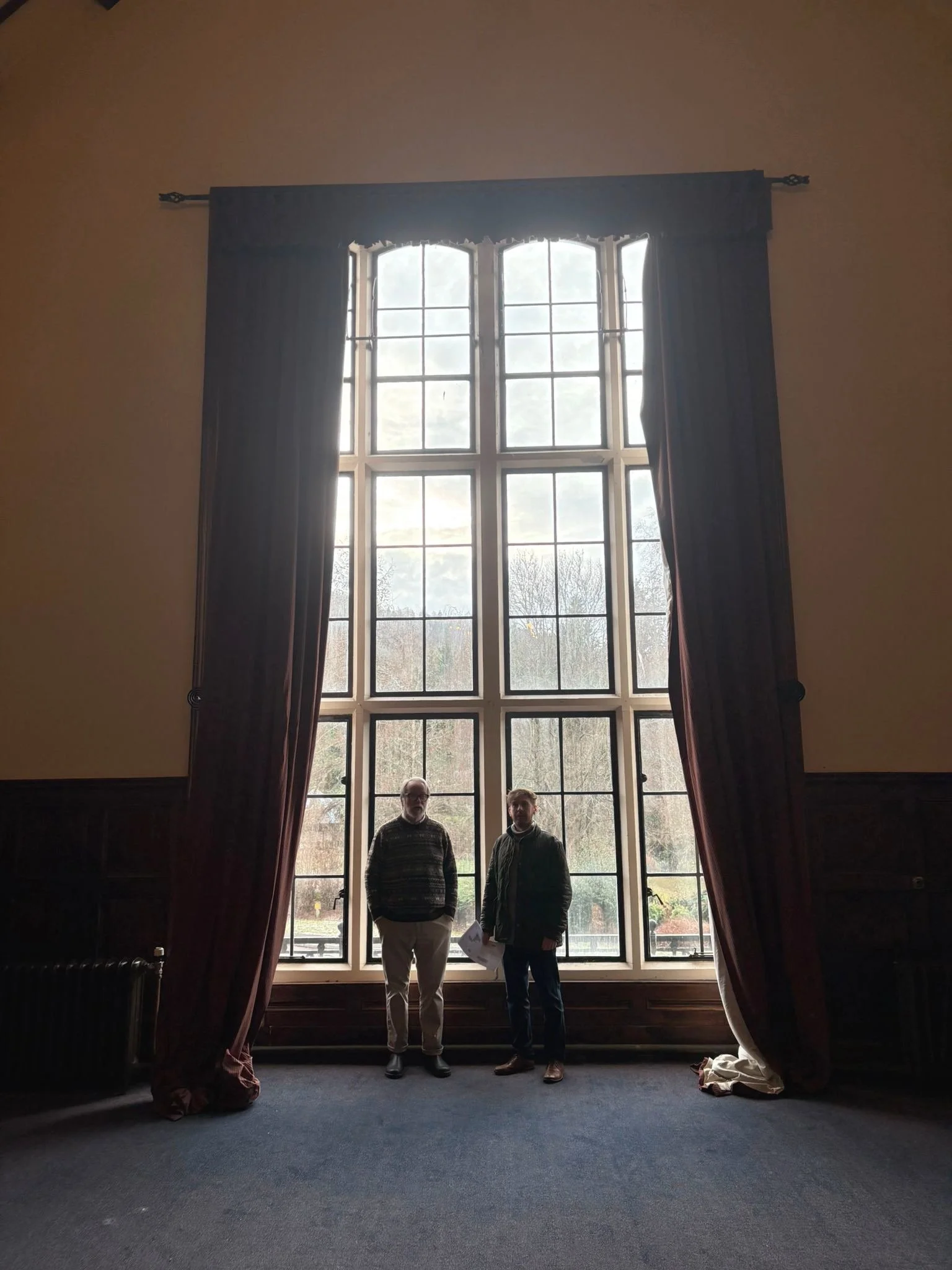 Two men standing in front of a large window with divided panes, tall curtains on either side, in a room with dark wood paneling and carpet.