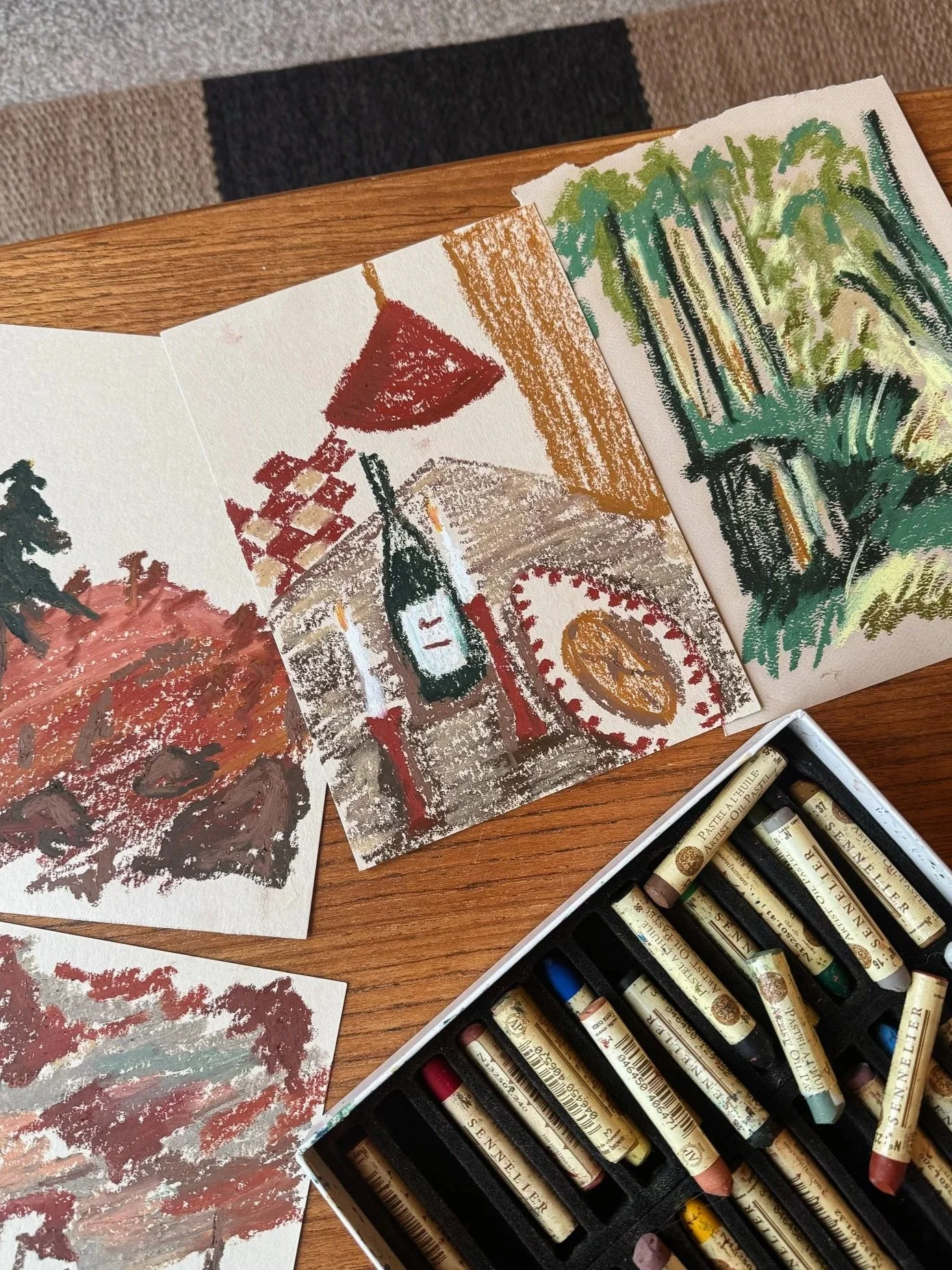 Colorful chalk drawings on paper depicting a landscape and a still life with a wine bottle, a red umbrella, and a plate of food, with a box of soft pastels nearby.
