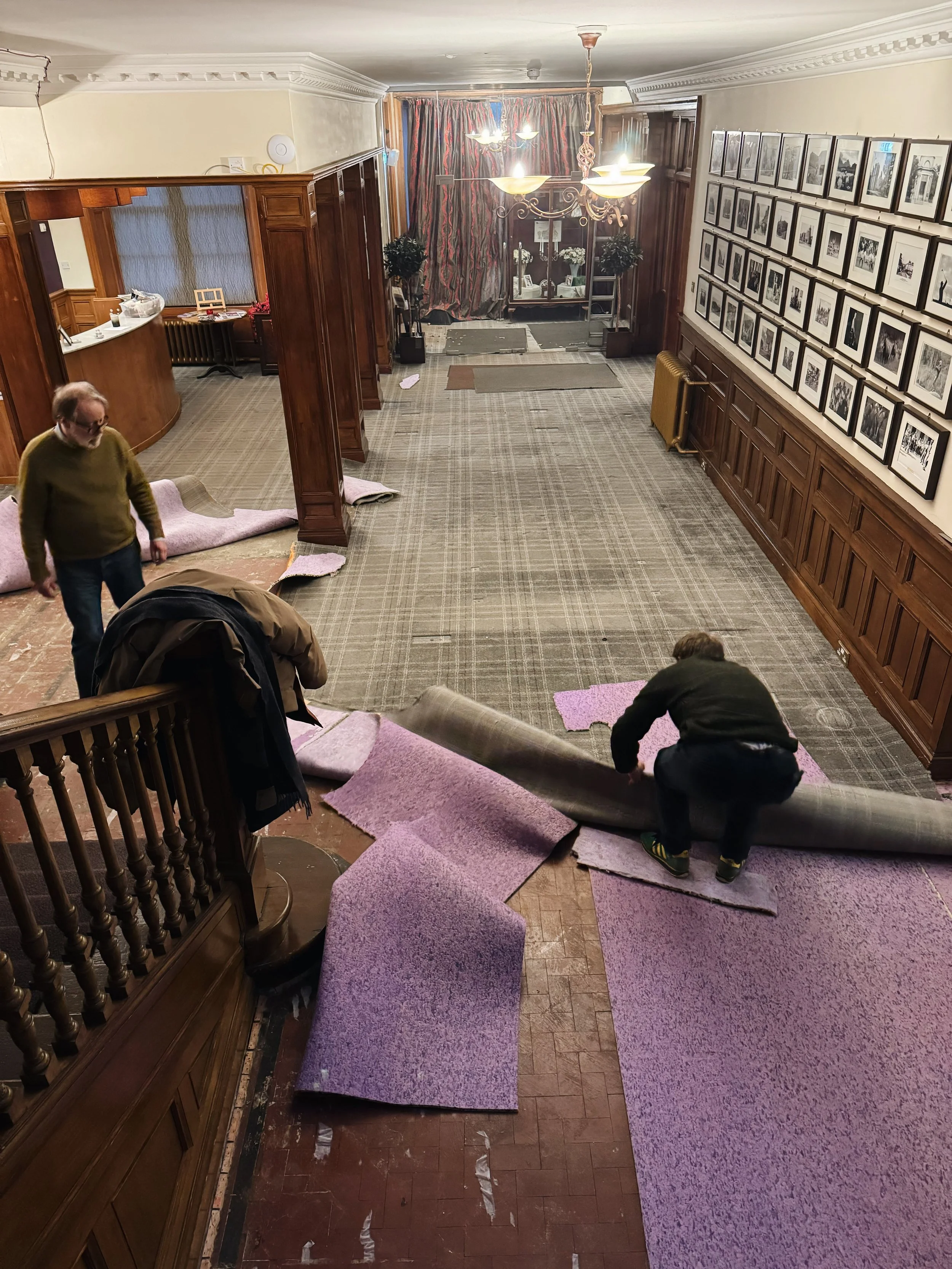 People are laying down and removing purple carpets in a large, well-lit room with framed photos on the wall, a chandelier, and wooden paneling.