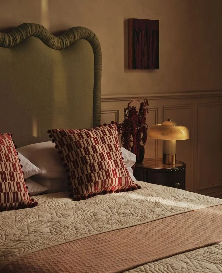 A cozy bedroom with a sage green upholstered headboard, two plaid pillows, a quilted bedspread, and a wooden nightstand with a vintage lamp and dried flowers in a vase.