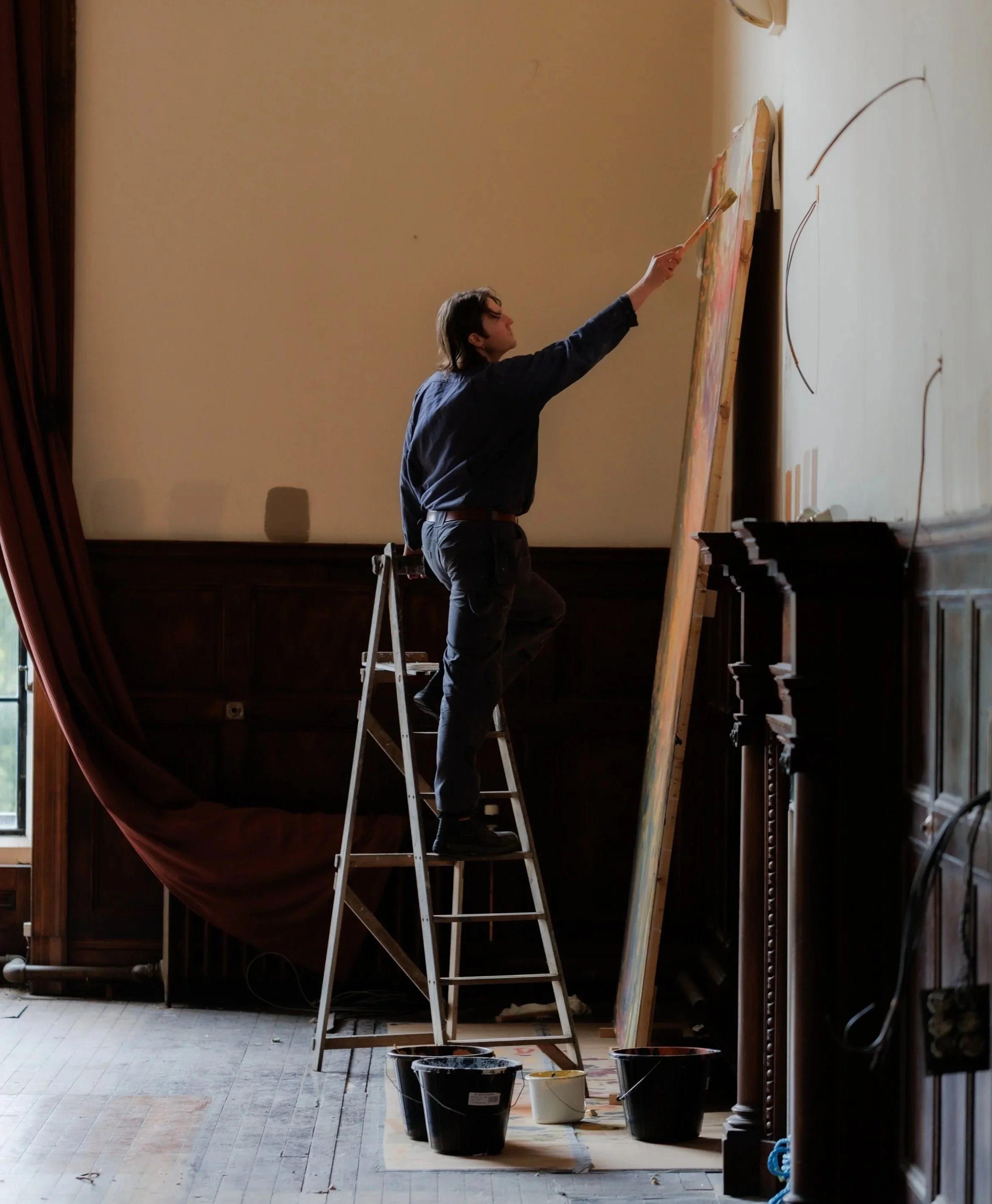 Painter felix zant in the birnam baronial hall. Person on a ladder painting on a large canvas in a room with wooden paneling.