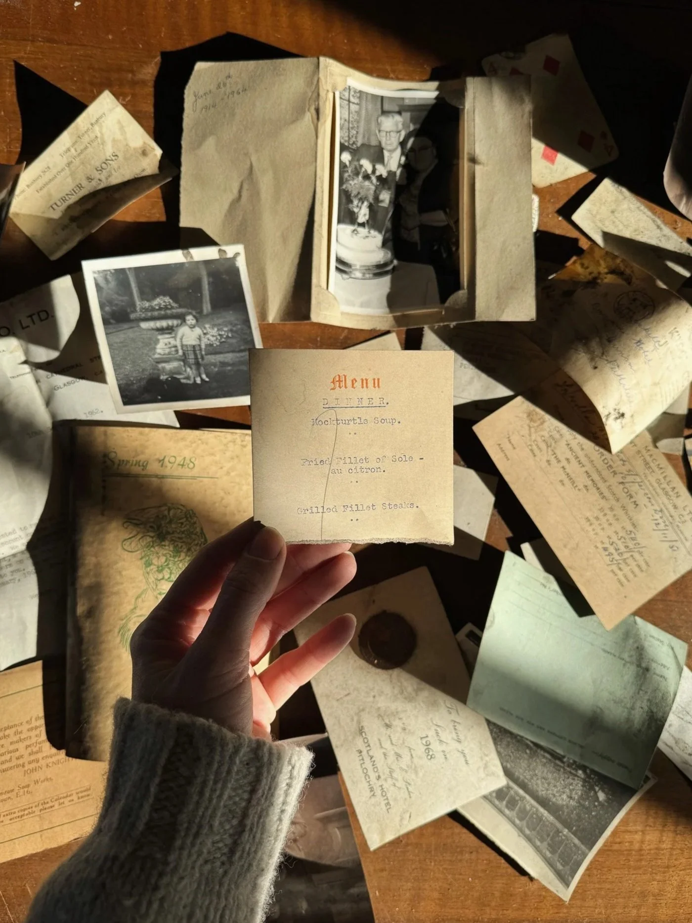 A hand holding a vintage menu from 1948 with dishes like mock turtle soup and grilled fillet steaks, surrounded by old photographs, papers, and a wooden table surface.