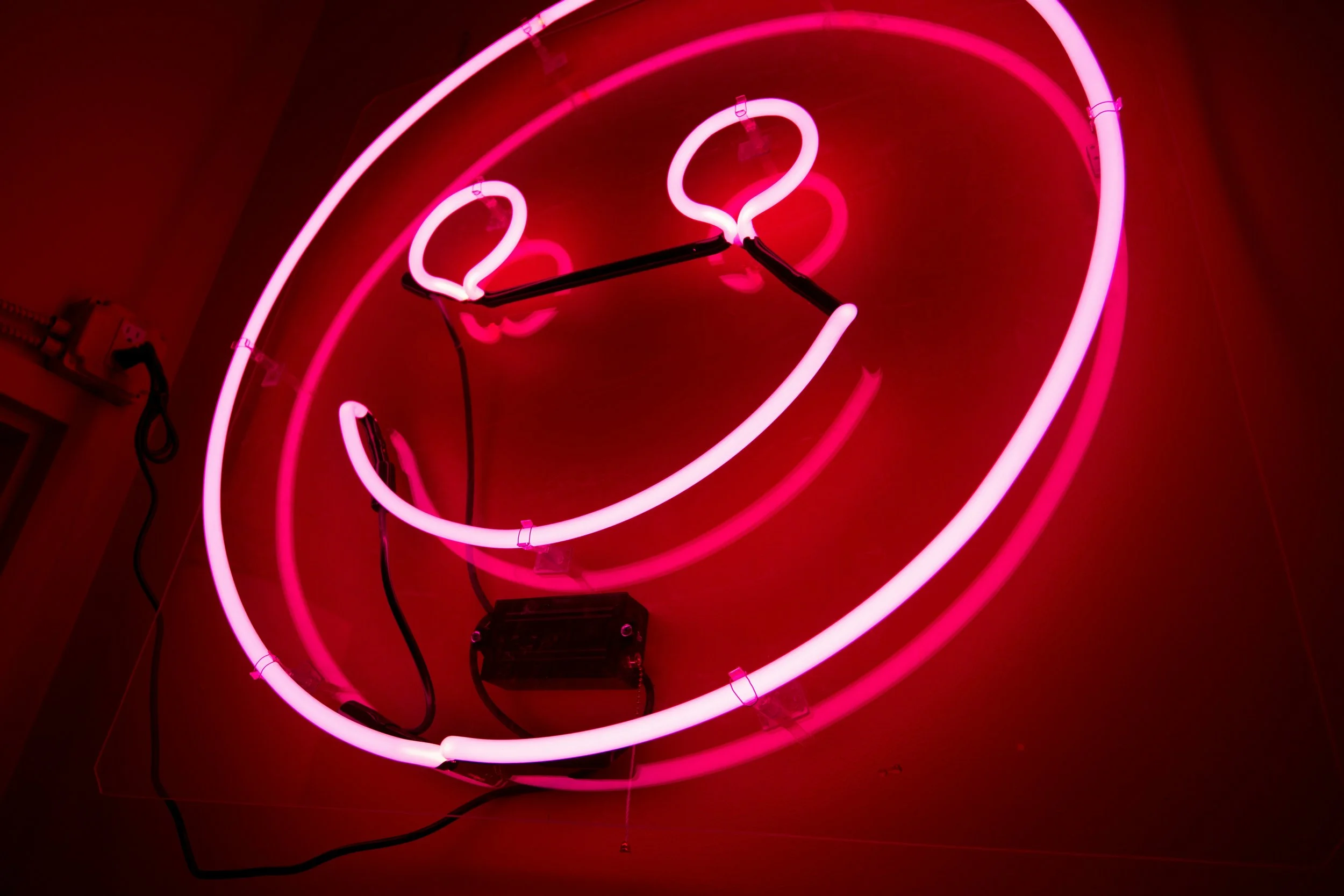A lit red-pink neon smiley face sign in a dark room