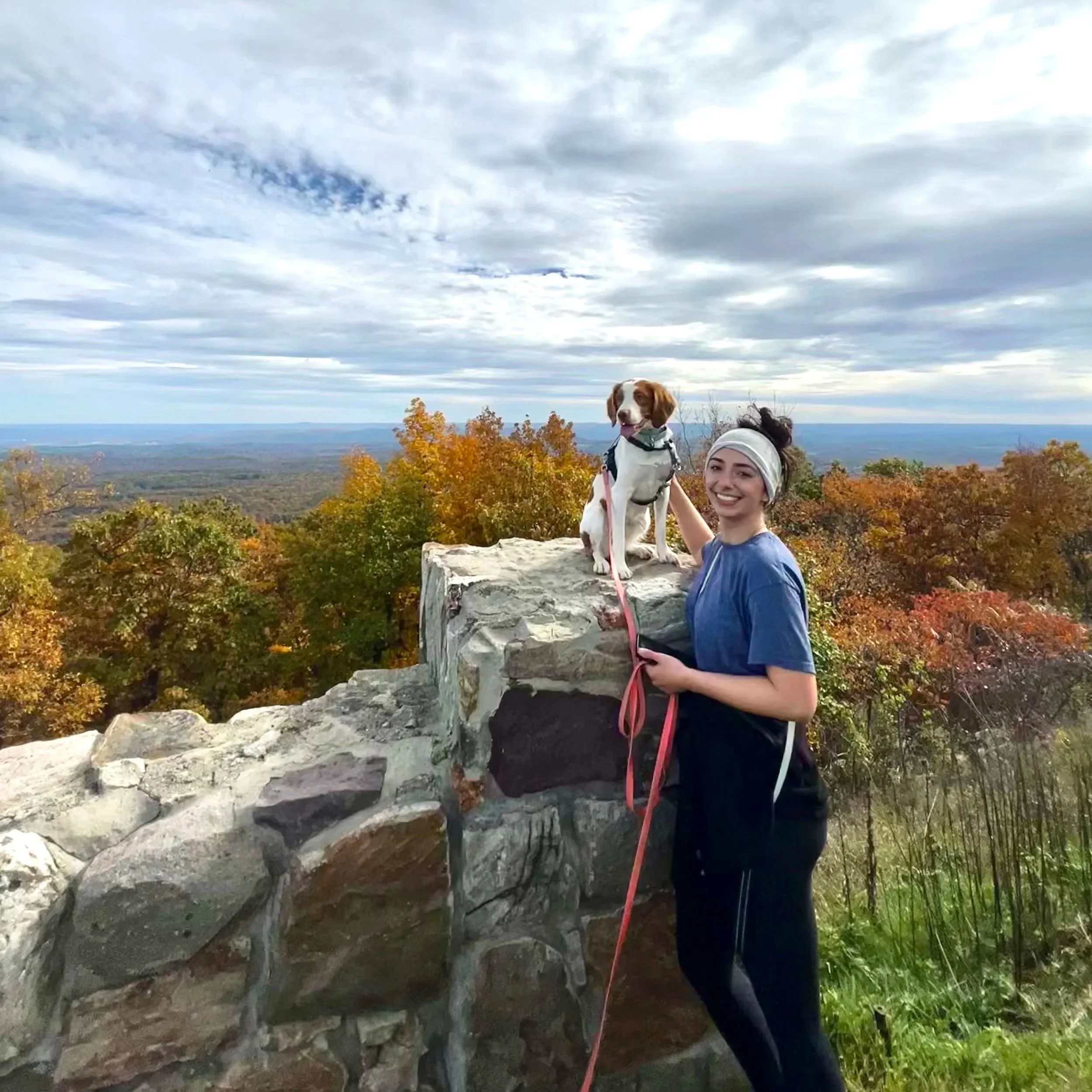 A young woman with a brittany spaniel holding a long leash posing for a photo after a hike 