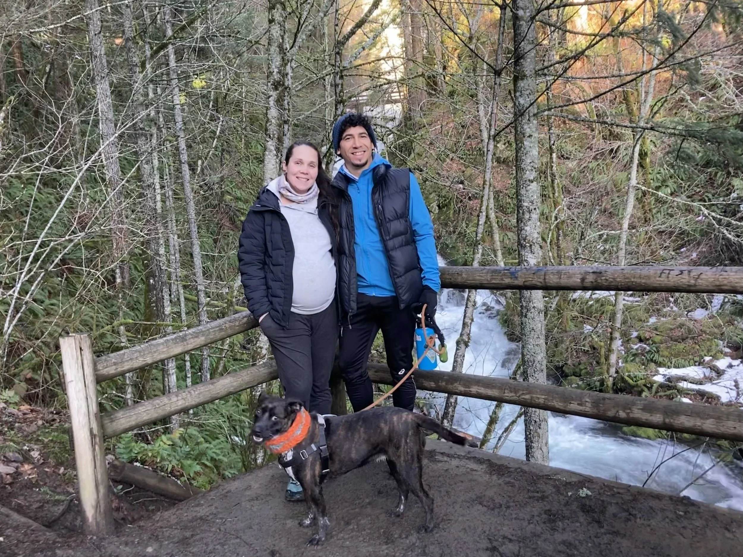 A man and pregnant woman smiling with a dog standing on a trail in a forest with trees and a river in the background.