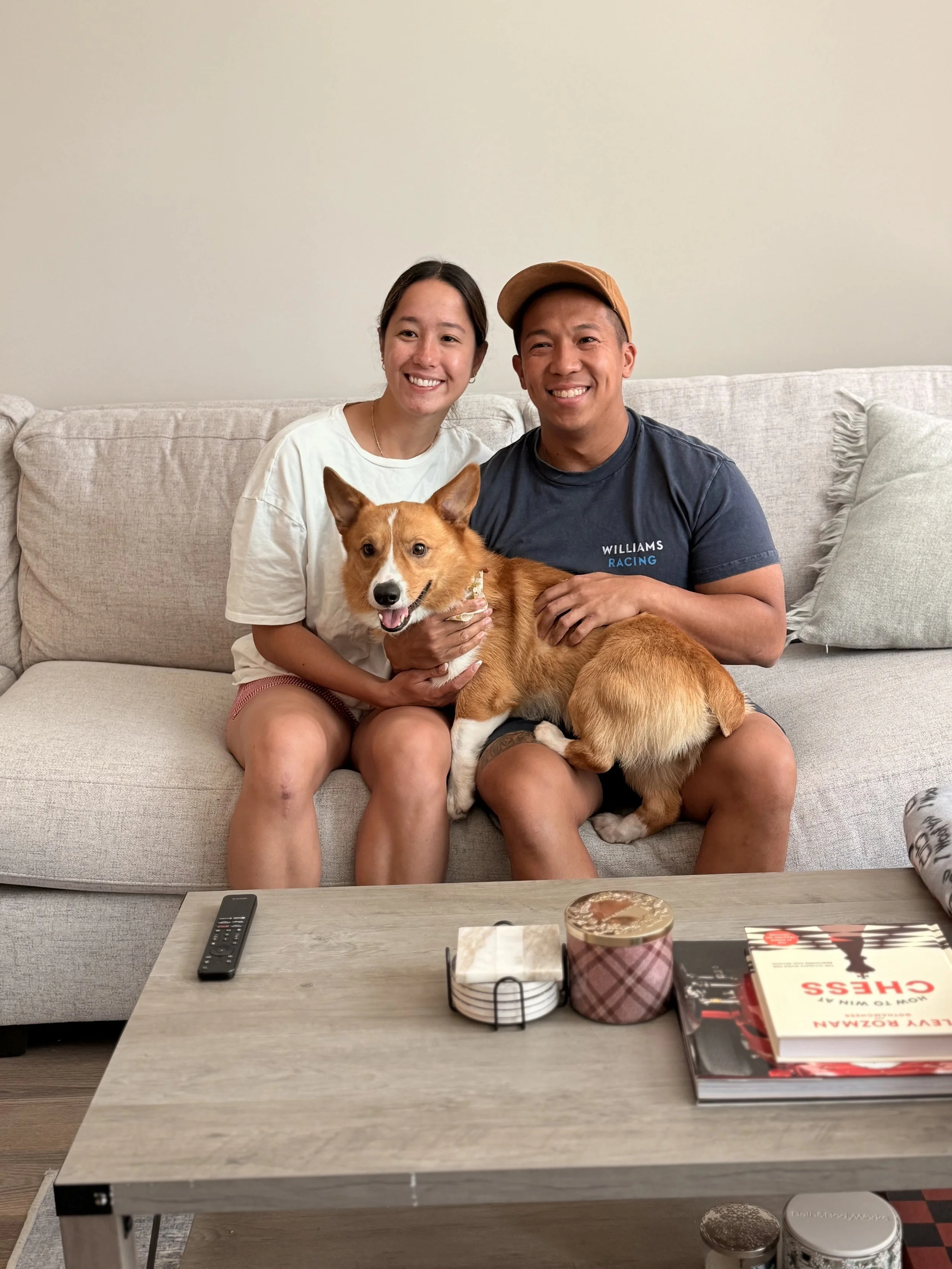 A smiling couple sitting on a beige couch holding a happy corgi between them in a cozy living room.