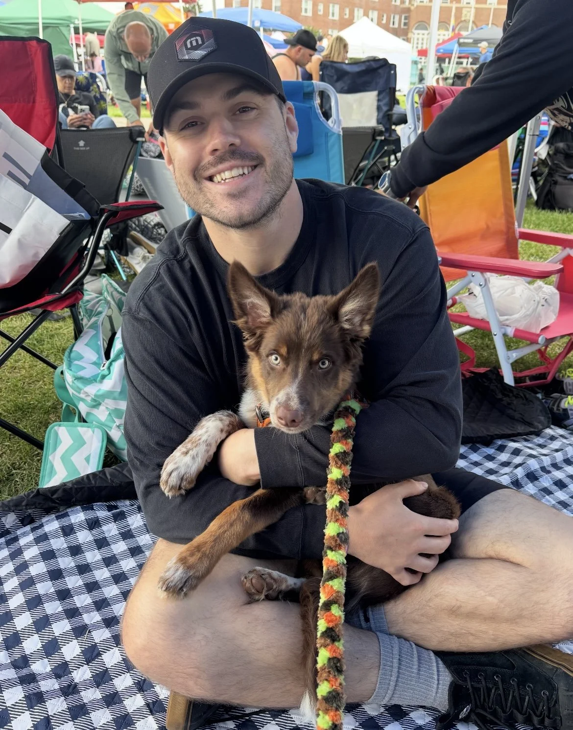 A man smiling while holding a border collie puppy at an outdoor event, sitting on a blanket with tents and people in the background.