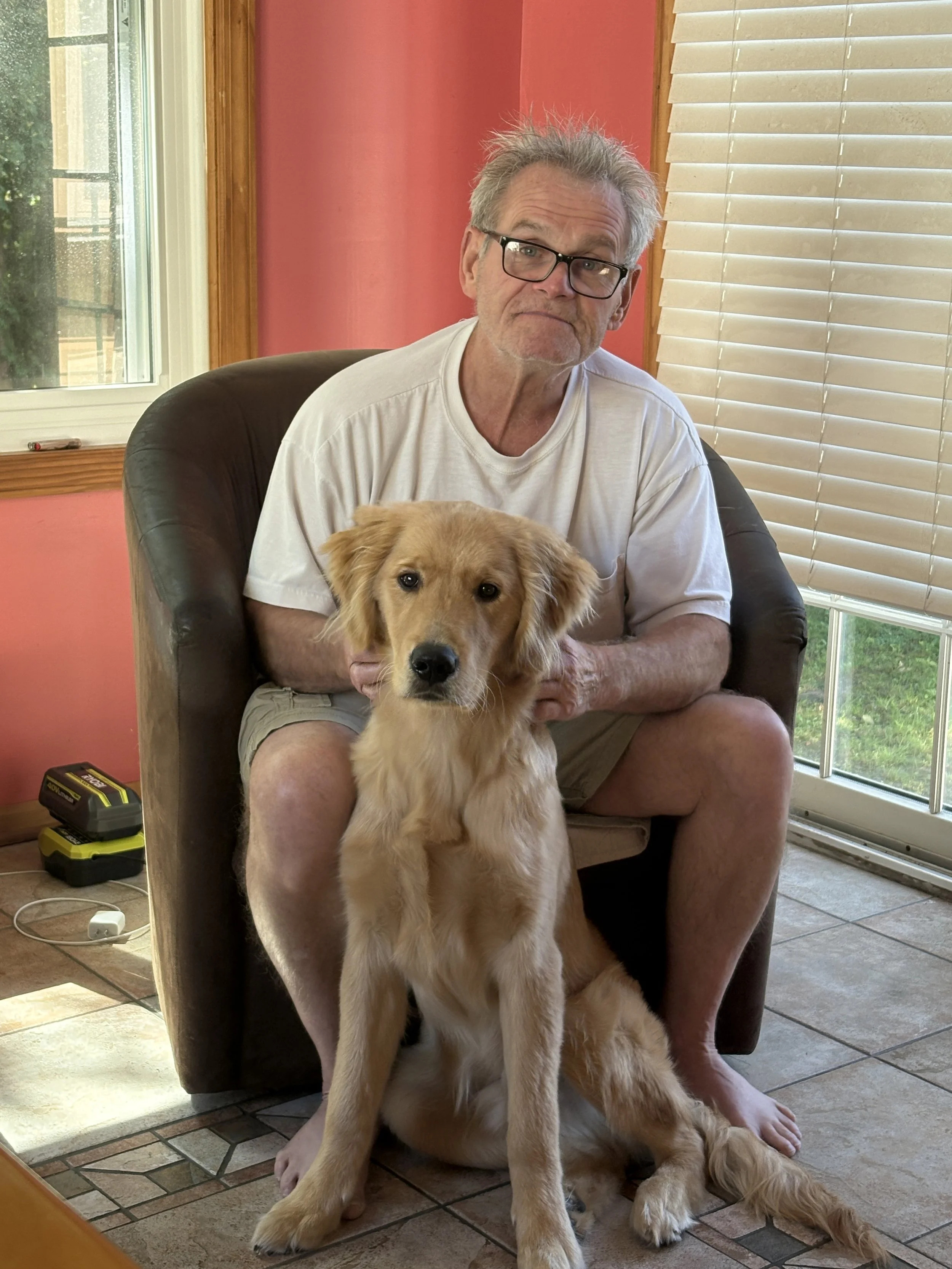 An elderly man with glasses sitting on a chair petting a golden retriever puppy