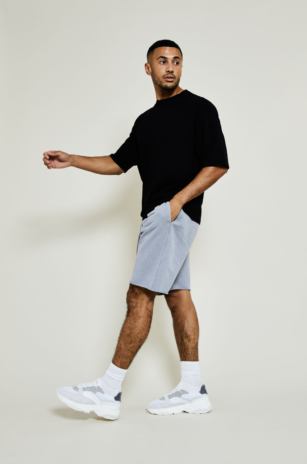 Young man standing against a plain light-colored background, wearing a loose black T-shirt, light gray shorts, white sneakers, and white socks, with one hand in his pocket and the other extended.