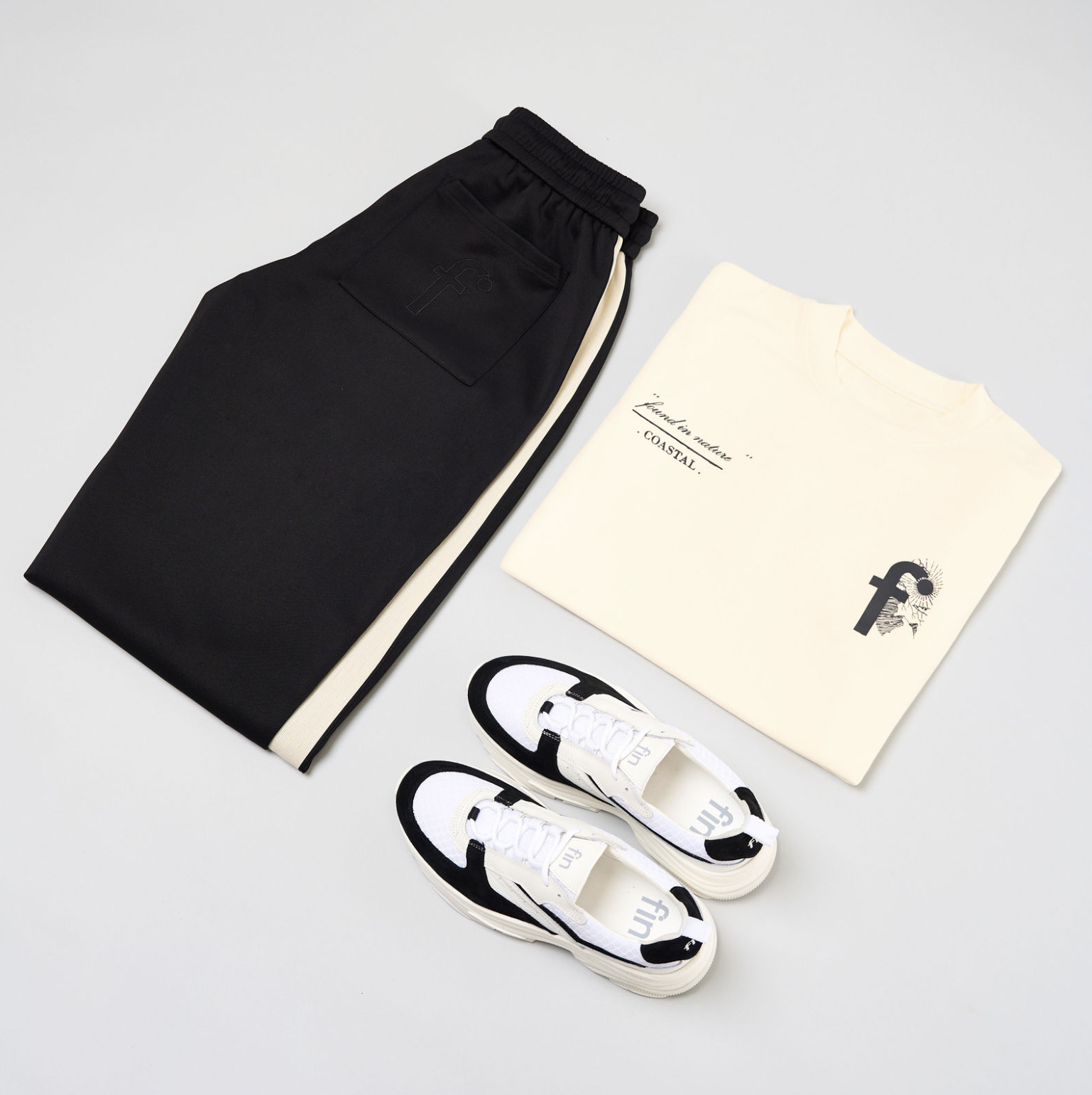 Black and white soccer-inspired sneakers, cream-colored t-shirt with black text, and black shorts with a pocket, arranged on a white surface.