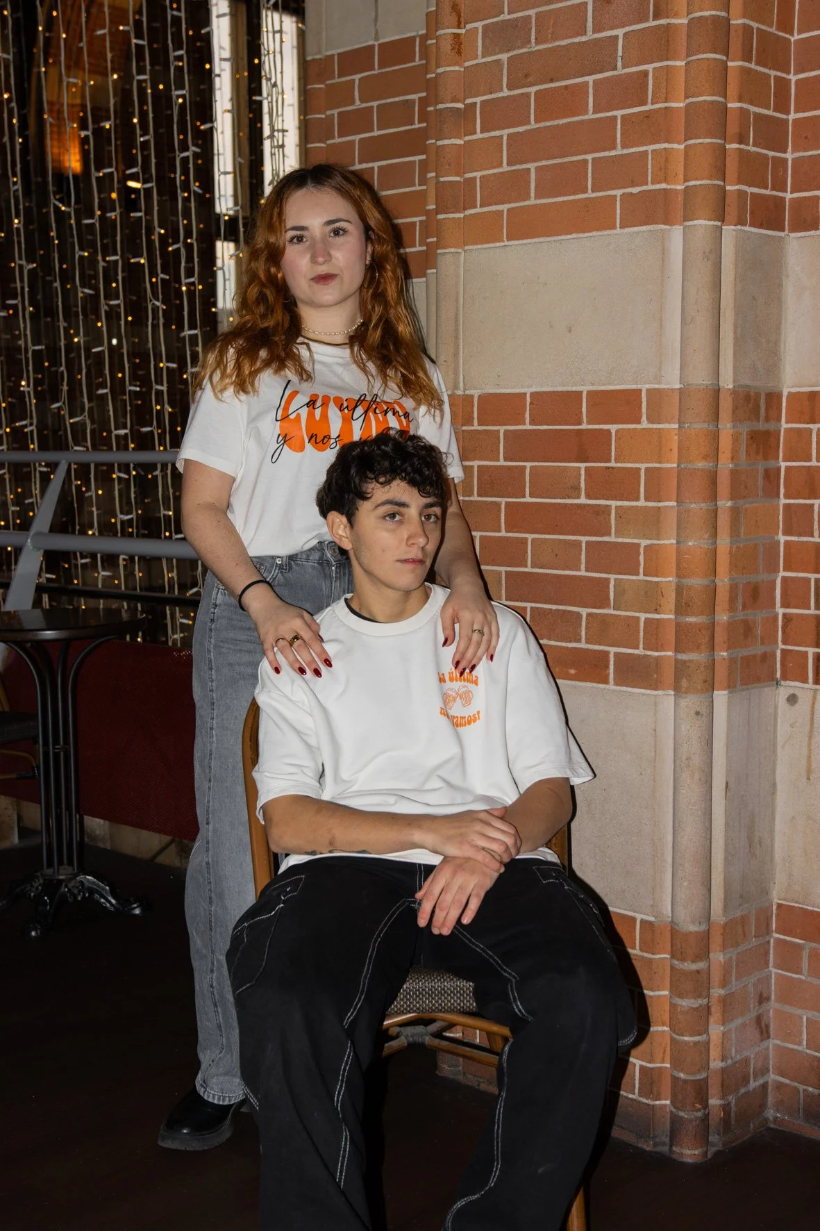 Dos jóvenes, uno sentado y otro de pie, con camisetas blancas con texto en rojo, en un lugar con paredes de ladrillos. La mujer de cabello rizado y mirada seria, y el joven con cabello rizado negro, observando hacia delante.