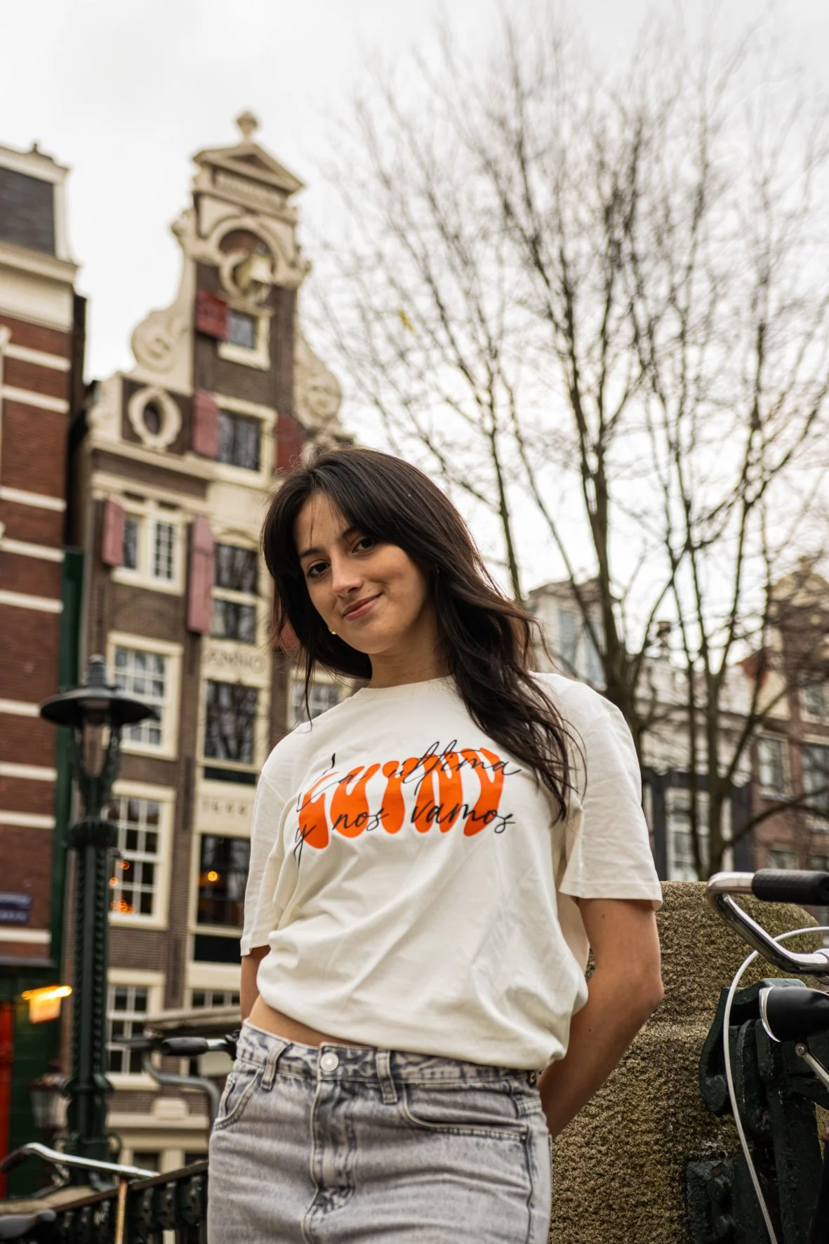 Mujer sonriendo con camiseta blanca y pantalones de mezclilla en calle con edificios históricos y árboles desnudos.