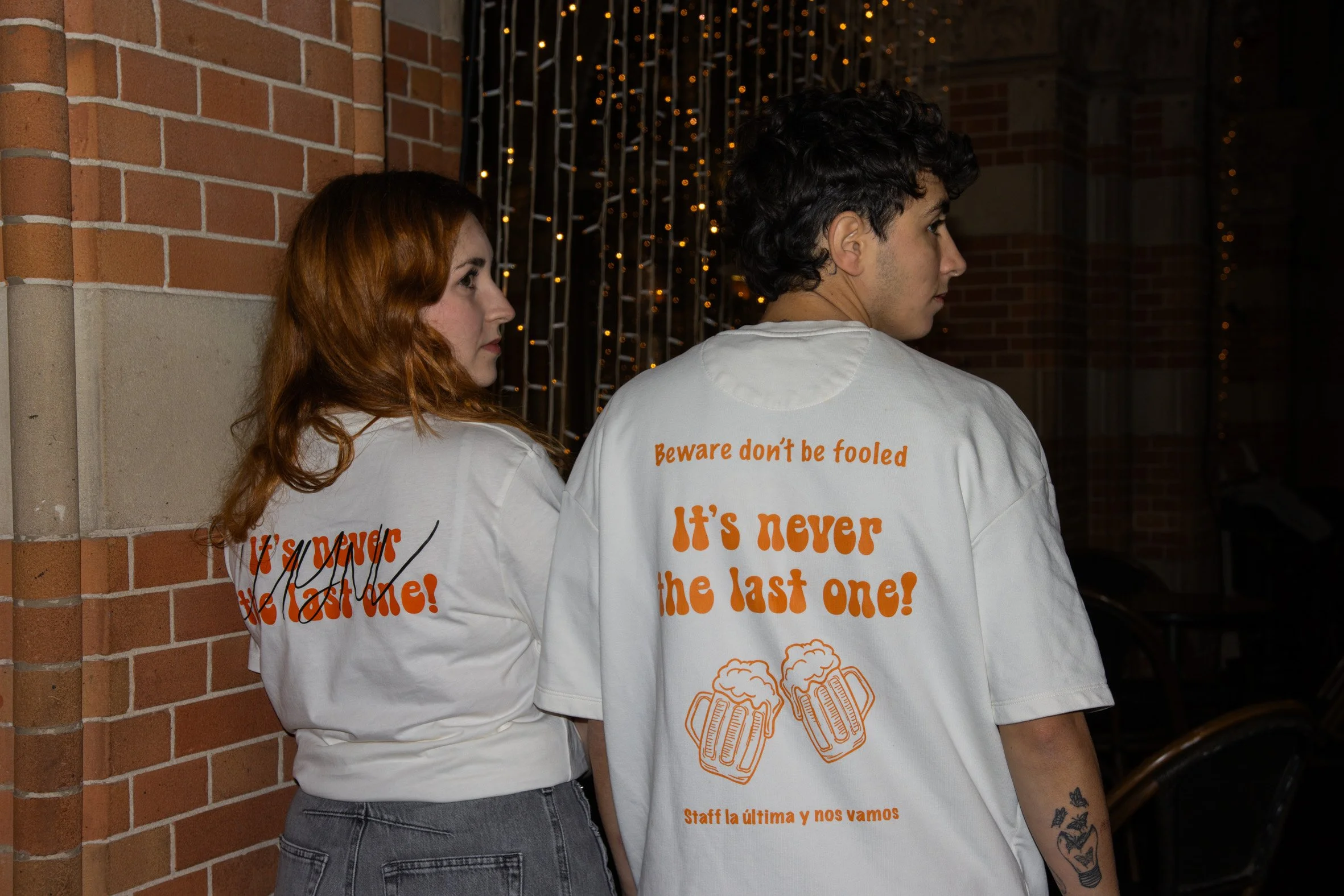Dos personas con camisetas blancas con texto en inglés y dibujos de jarras de cerveza, de espaldas, en un entorno con paredes de ladrillo y luces decorativas.