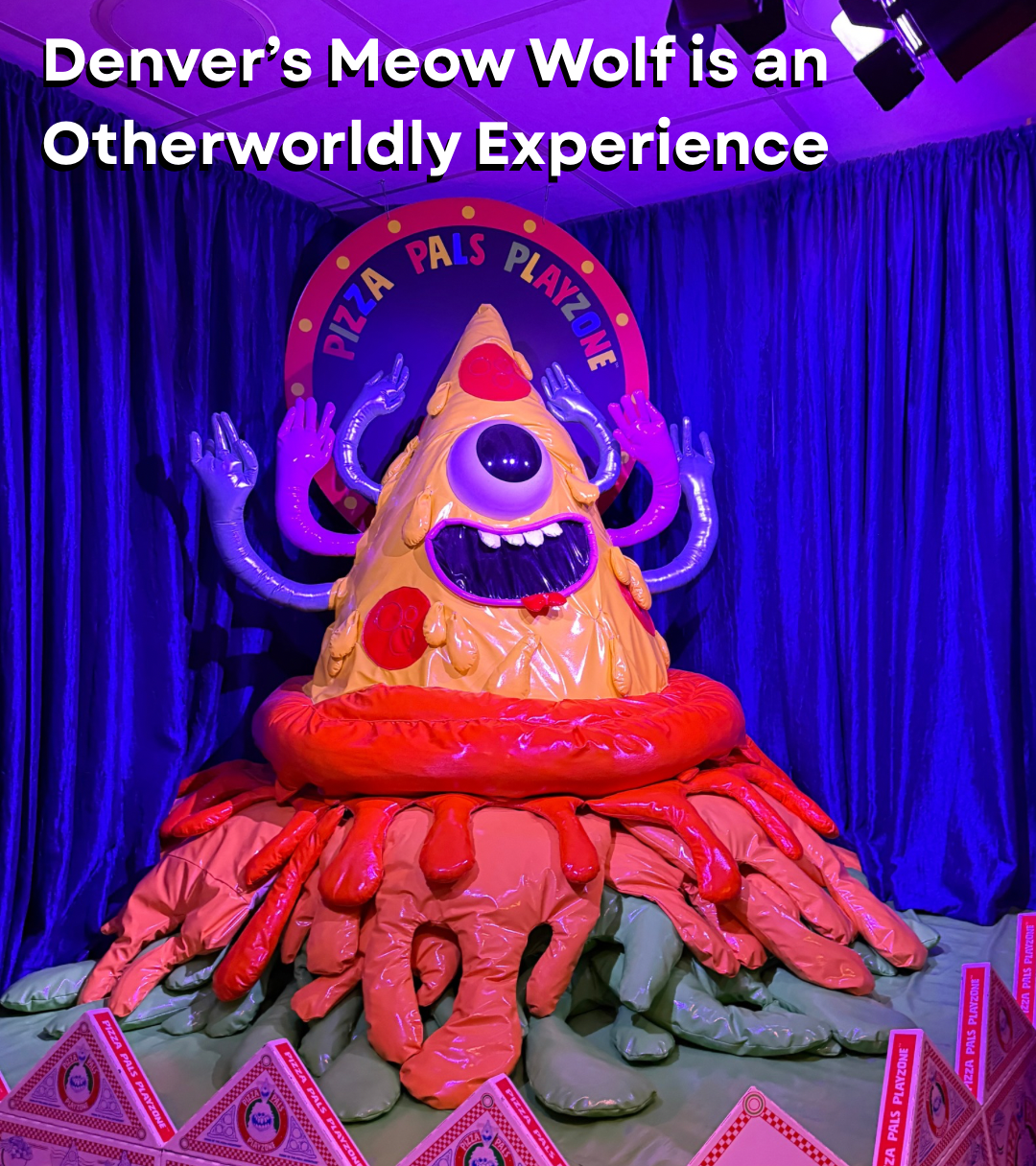 Colorful sculpture of a one-eyed monster with purple arms and a pizza body, surrounded by a pizza-themed backdrop and blue curtains at Denver's Meow Wolf. 