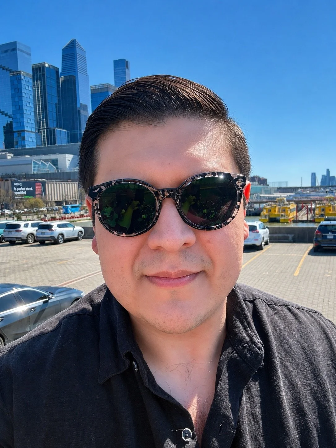 A man wearing sunglasses taking a selfie in a parking lot with city buildings, cars, and yellow water taxis in the background on a sunny day.