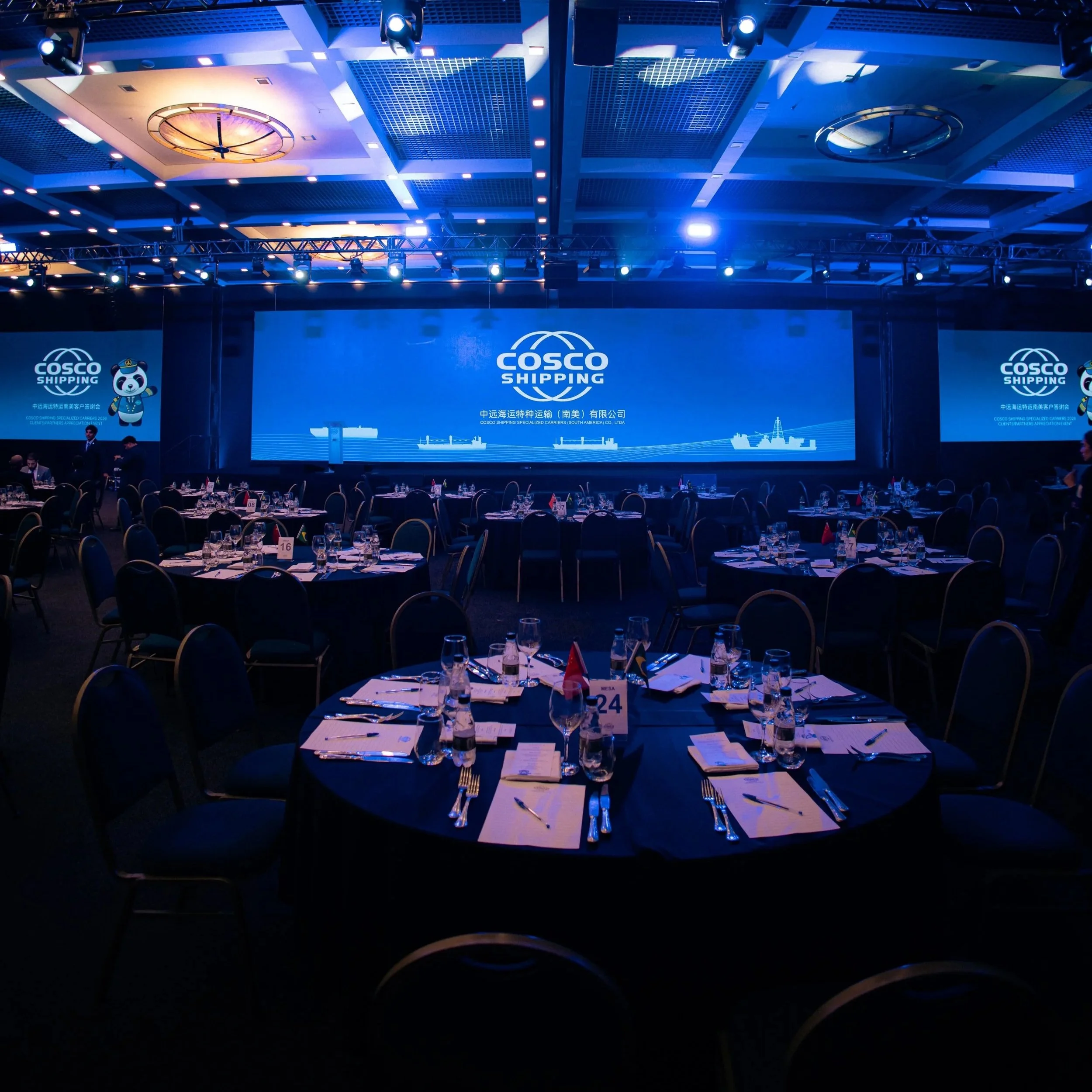 Grande sala de conferências decorada com iluminação azul e mesas arrumadas com utensílios para jantar, em um evento da Cosco Shipping, com uma grande tela de apresentação ao fundo exibindo o logo da empresa e temas relacionados à logística marítima.