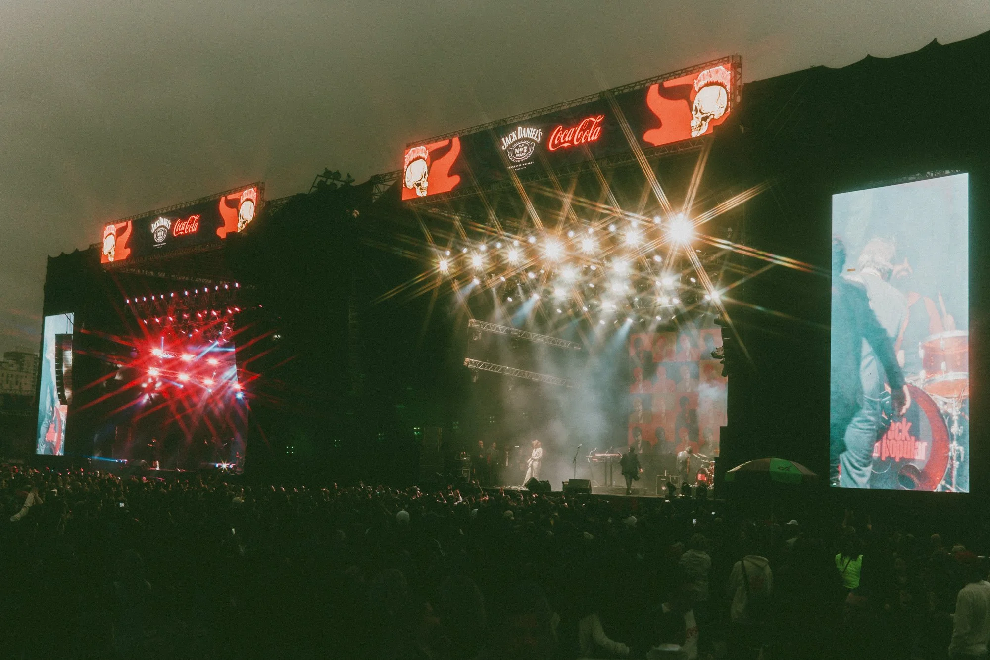 Imagem de um grande show ao ar livre à noite com palco iluminado, telas de vídeo ao lado, luzes brilhantes e multidão assistindo ao espetáculo. ativação de marca coca-cola jack daniels