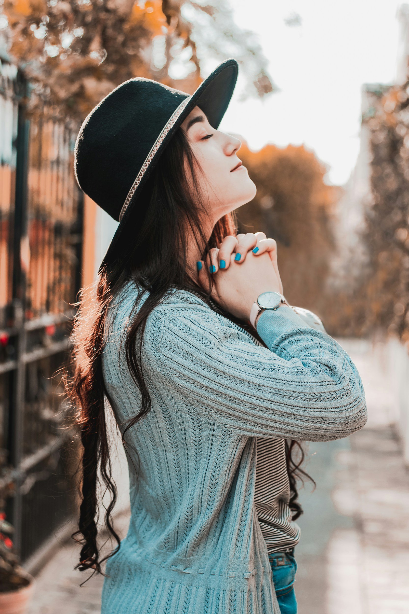 A perfectionist with a light blue cable-knit sweater, and a watch on her wrist, stands outdoors with her eyes closed and hands clasped near her chin. She is in profile against a blurred background of trees with orange leaves and a street scene.