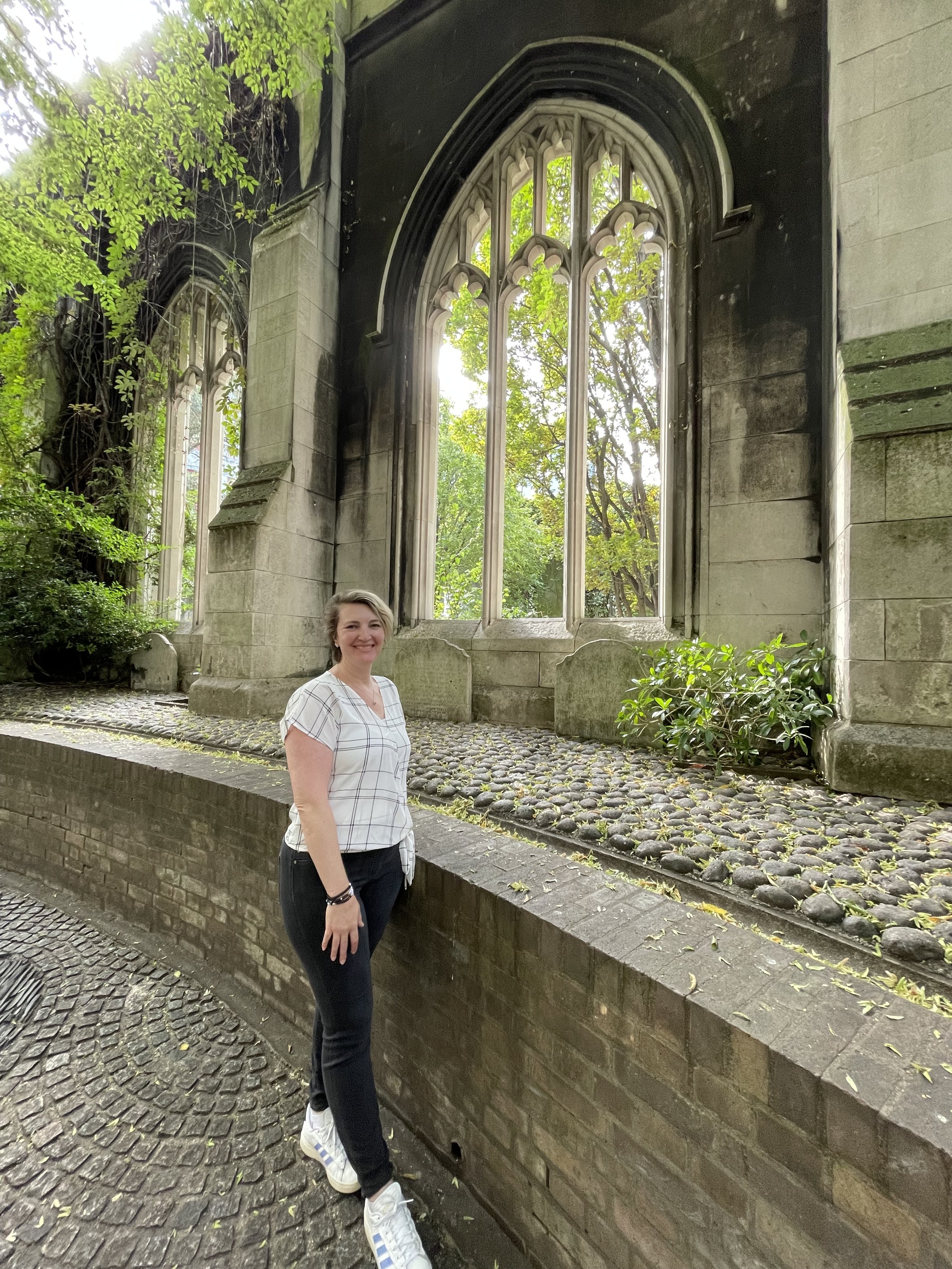 A woman in front of ruins of a church in London with the sun shining and greenery around.