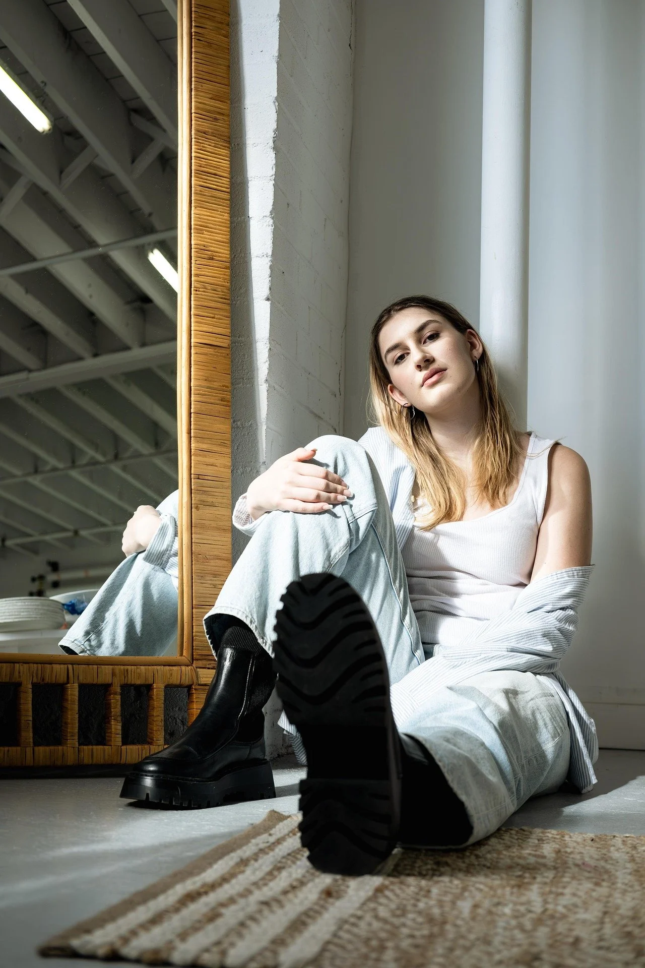 A perfectionist teen sitting on the floor near a mirror, wearing a white tank top, loose pants, and black boots, with a relaxed expression in an industrial-style room.