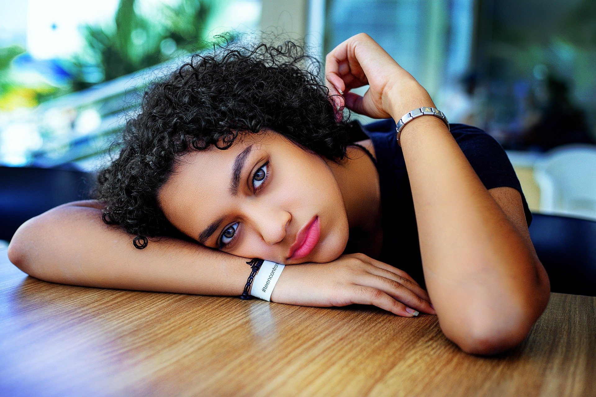 An anxious young woman with curly dark hair and blue eyes lying on a wooden table, resting her head on her arms, with a blurred background showing natural light and greenery.