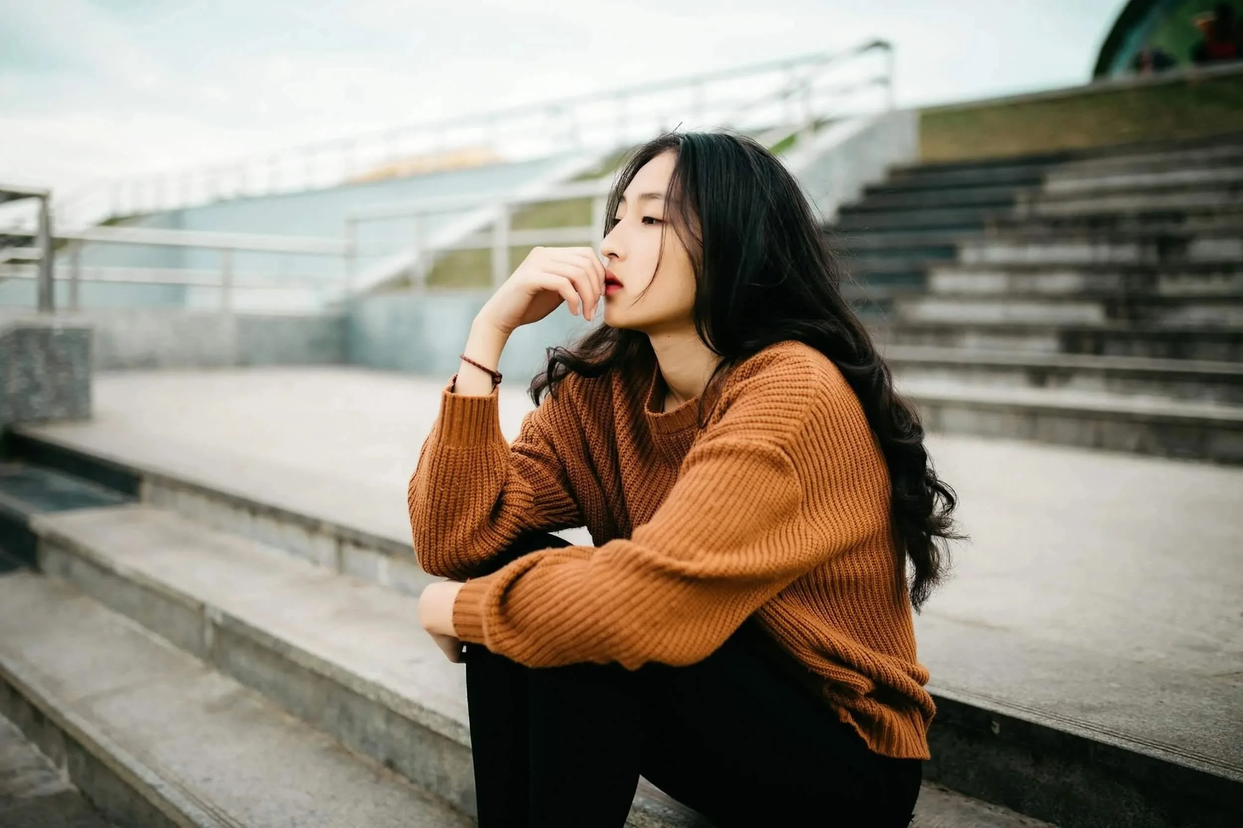 A  perfectionist young woman with long dark hair, wearing a brown sweater, sitting on concrete steps outdoors, with her hand near her face, looking contemplative.