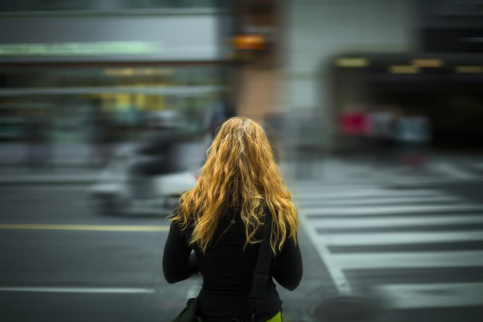 A worried woman with long, curly blonde hair standing at a crosswalk in an urban setting, with motion-blurred cars passing by.
