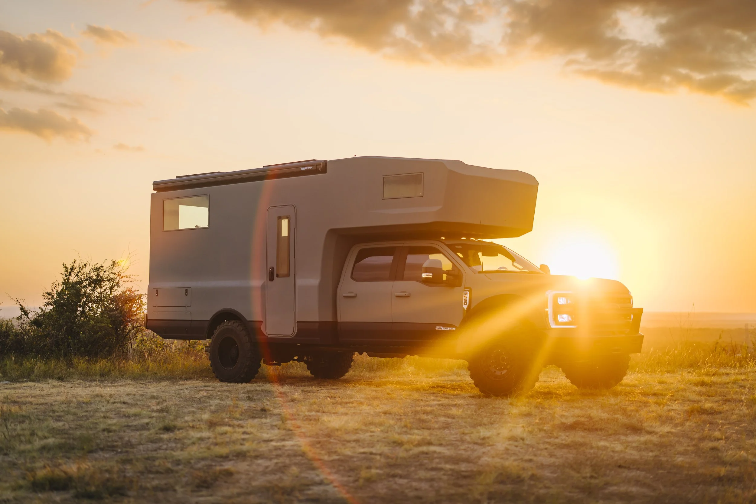 A large off-road vehicle with an attached camper traveling across open land at sunset.