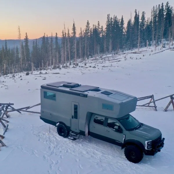 A gray camper van with an overhead living area parked on snow in a forested area at sunset.